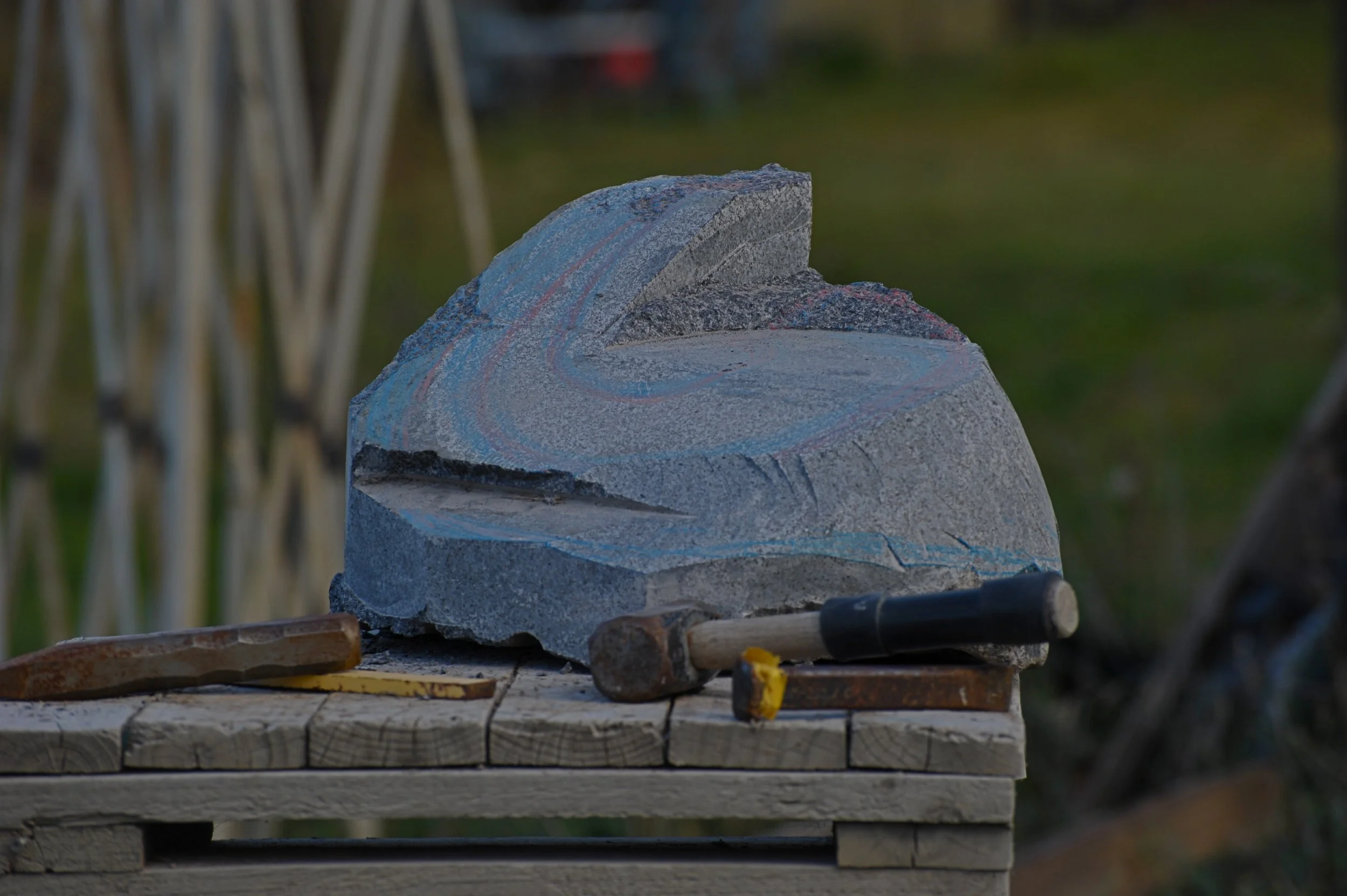 A roughly hewn stone with colorful chalk markings rests on a wooden pallet outdoors, with blurred grassy background and tools nearby.