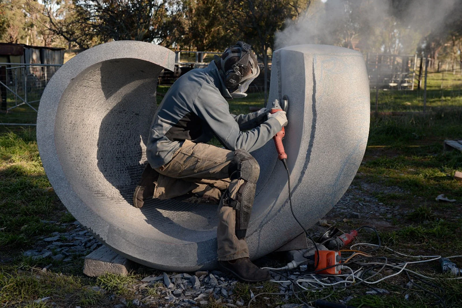 A person wearing a helmet and protective gear is using a power tool to carve a large, abstract stone sculpture outdoors during the day. The sculpture has a curved, hollow shape with a textured surface.