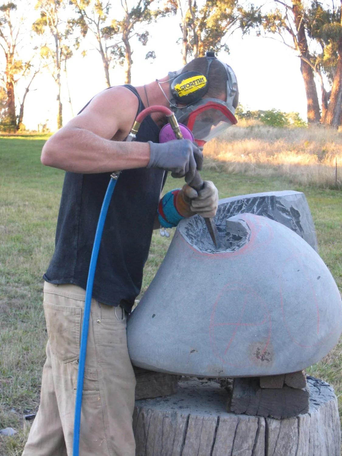 A person wearing safety gear, including a face mask, ear protection, and gloves, is carving a sculpture out of a large piece of stone outdoors in a grassy area with trees in the background.