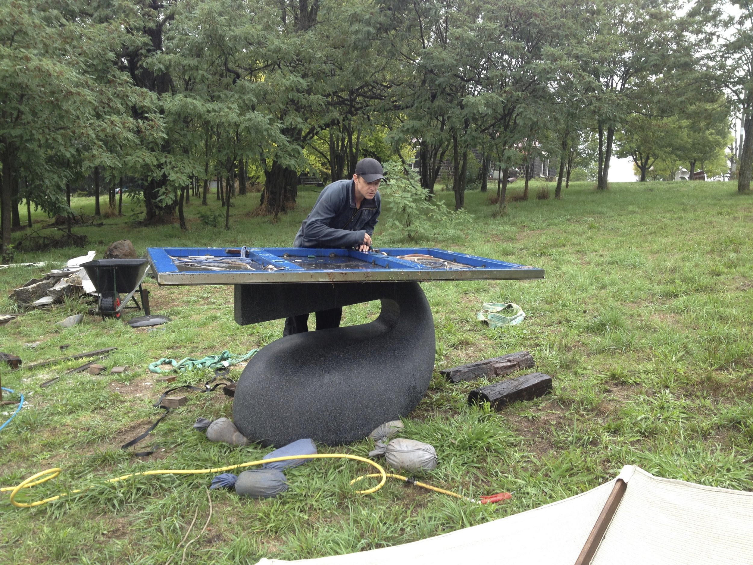 A man wearing a black cap and jacket is working outdoors at a table with various equipment, surrounded by trees and grassy area.
