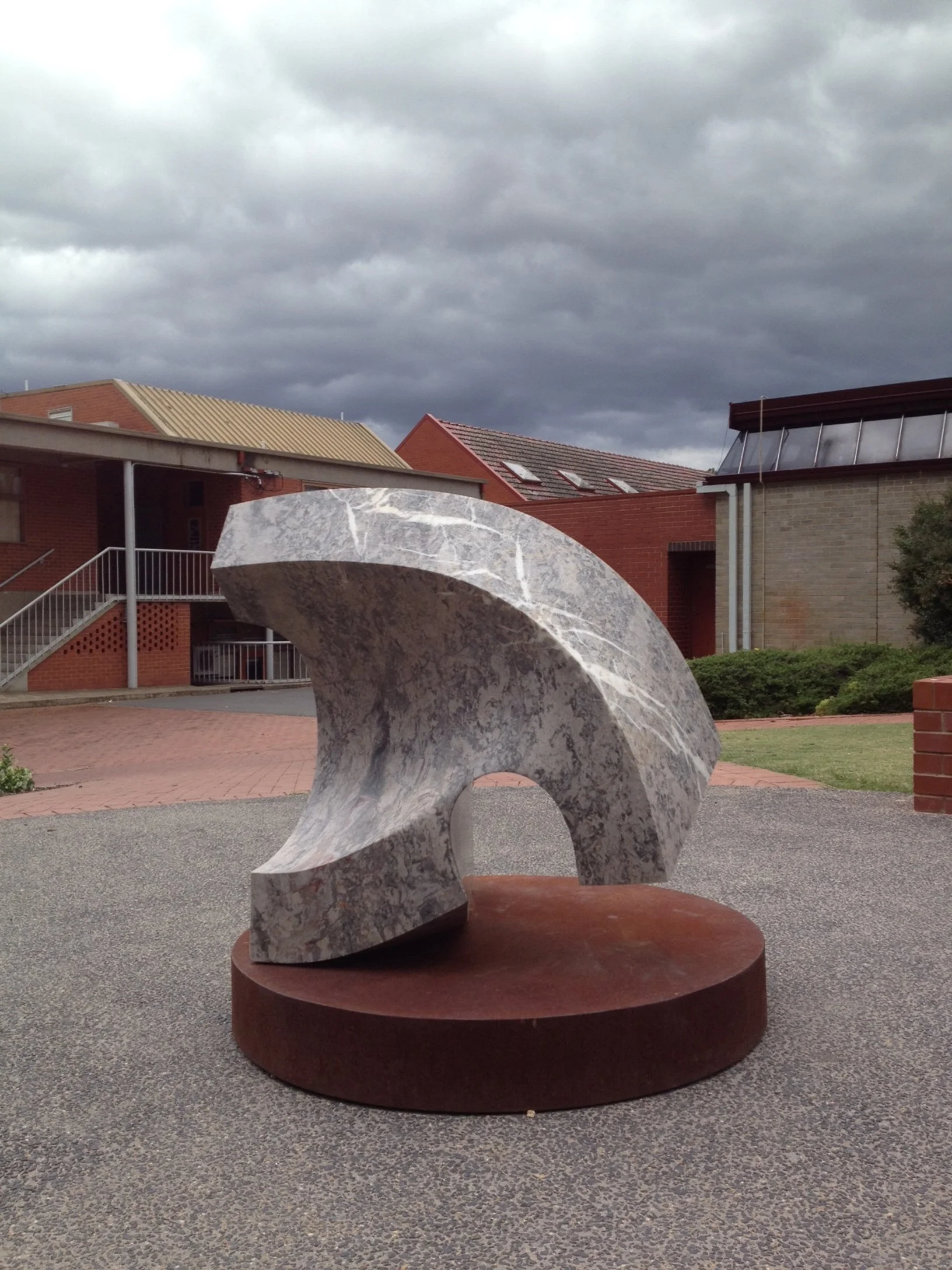 Abstract marble sculpture on a round base in an outdoor courtyard, with buildings and dark cloudy sky in the background.