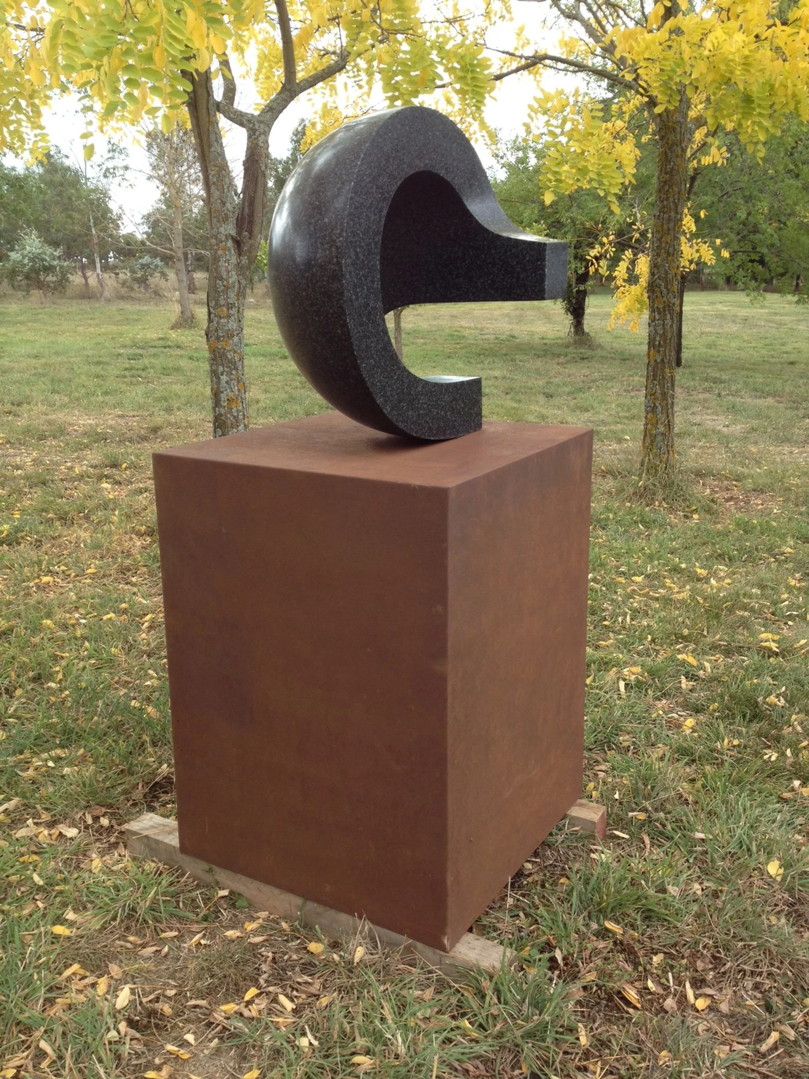 A large, black granite sculpture of the letter 'C' on a brown pedestal in an outdoor park with trees and grass.