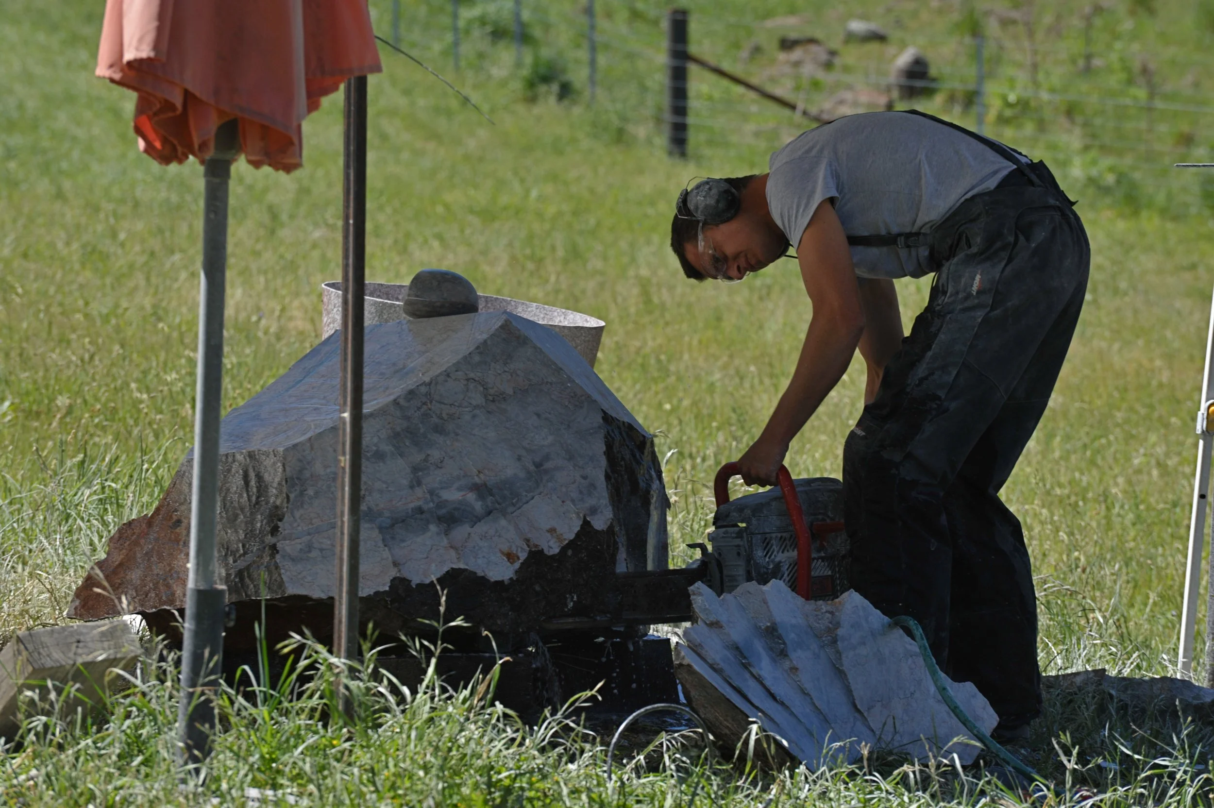 A man wearing safety goggles and a gray T-shirt is cutting a large piece of stone or rock outdoors with a power saw. There is green grass on the ground, and fencing and trees in the background.