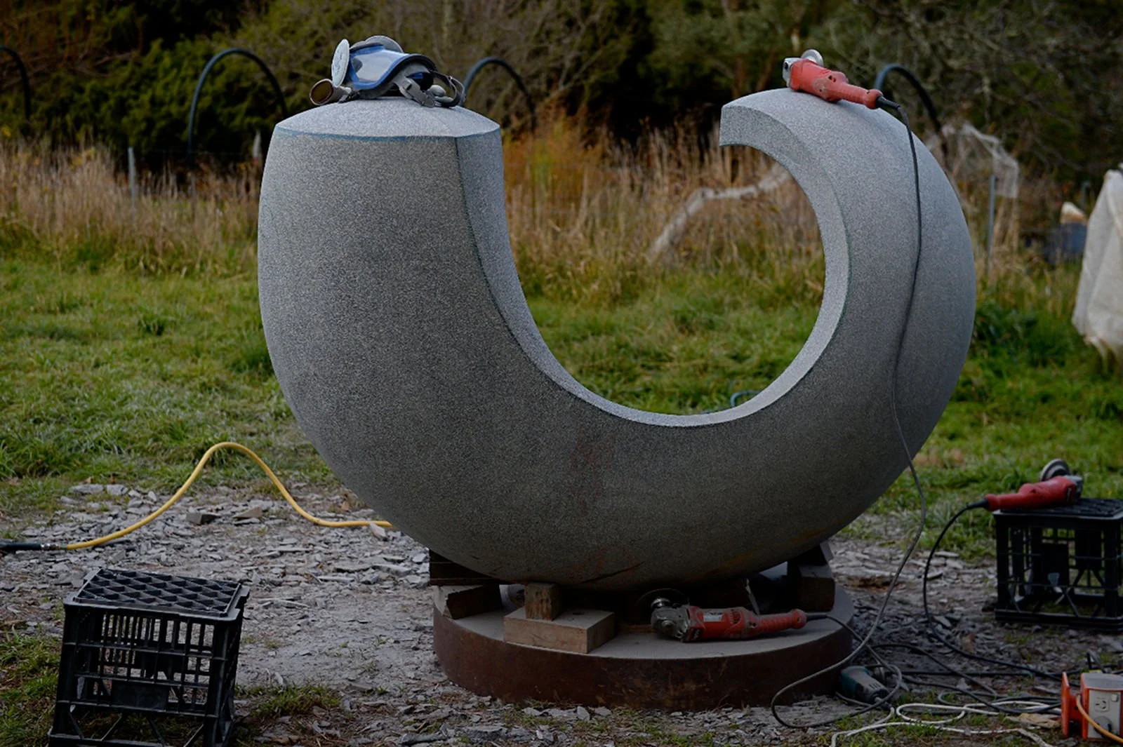 A large U-shaped stone sculpture outdoors, with construction tools, including a drill and an extension cord, surrounding it.