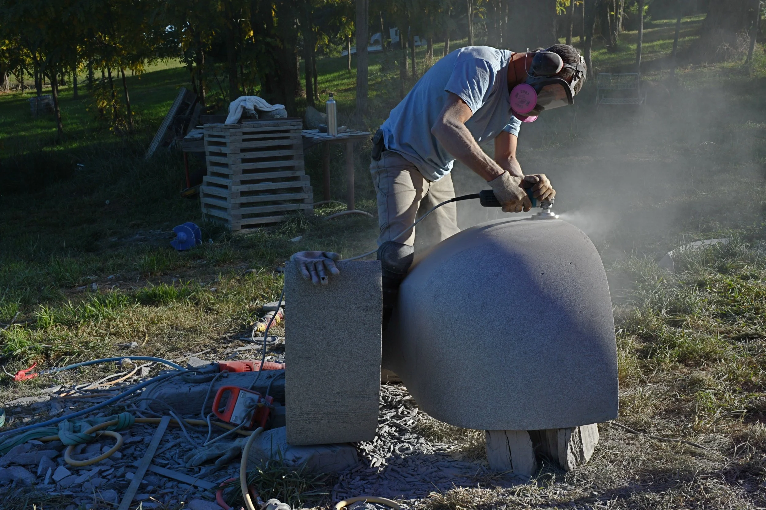 A man working outdoors with power tools on a large cylindrical object, wearing safety goggles, a face mask, gloves, and a blue shirt. There are trees and grass in the background, with various tools and materials scattered around.