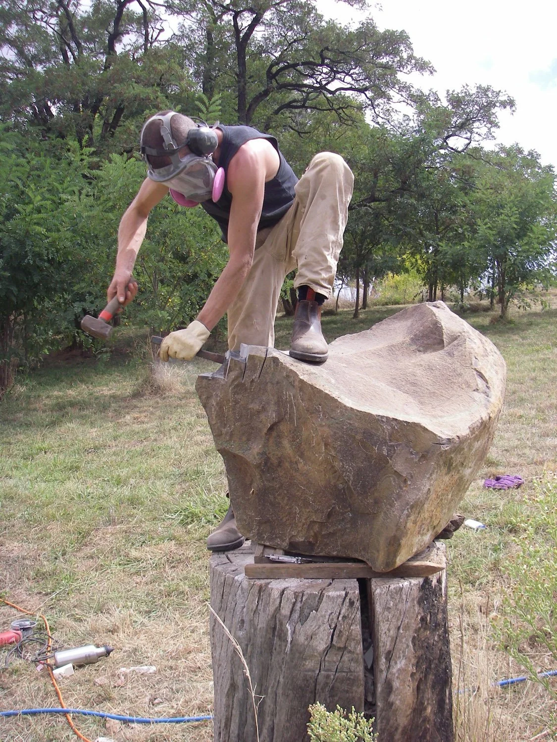 A man with safety goggles, mask, gloves, and boots is standing on a large rock, hammering on it outdoors on a tree stump, with trees and greenery in the background.