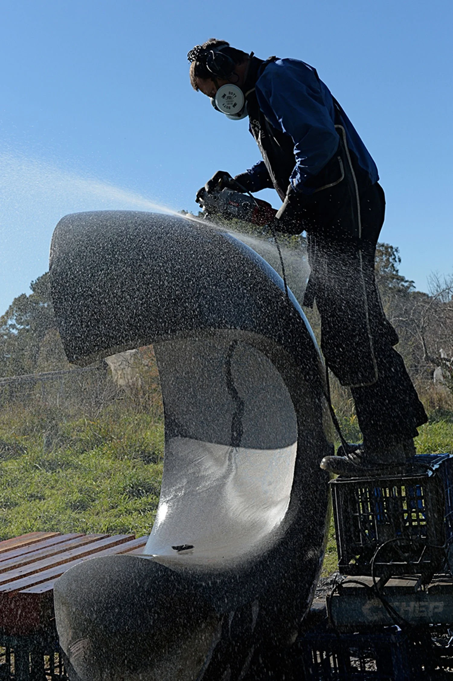 A person in a blue jacket and black pants wearing a mask, operating a grinder to cut a large piece of black granite, with water spraying from the cutting process on a sunny day outdoors.