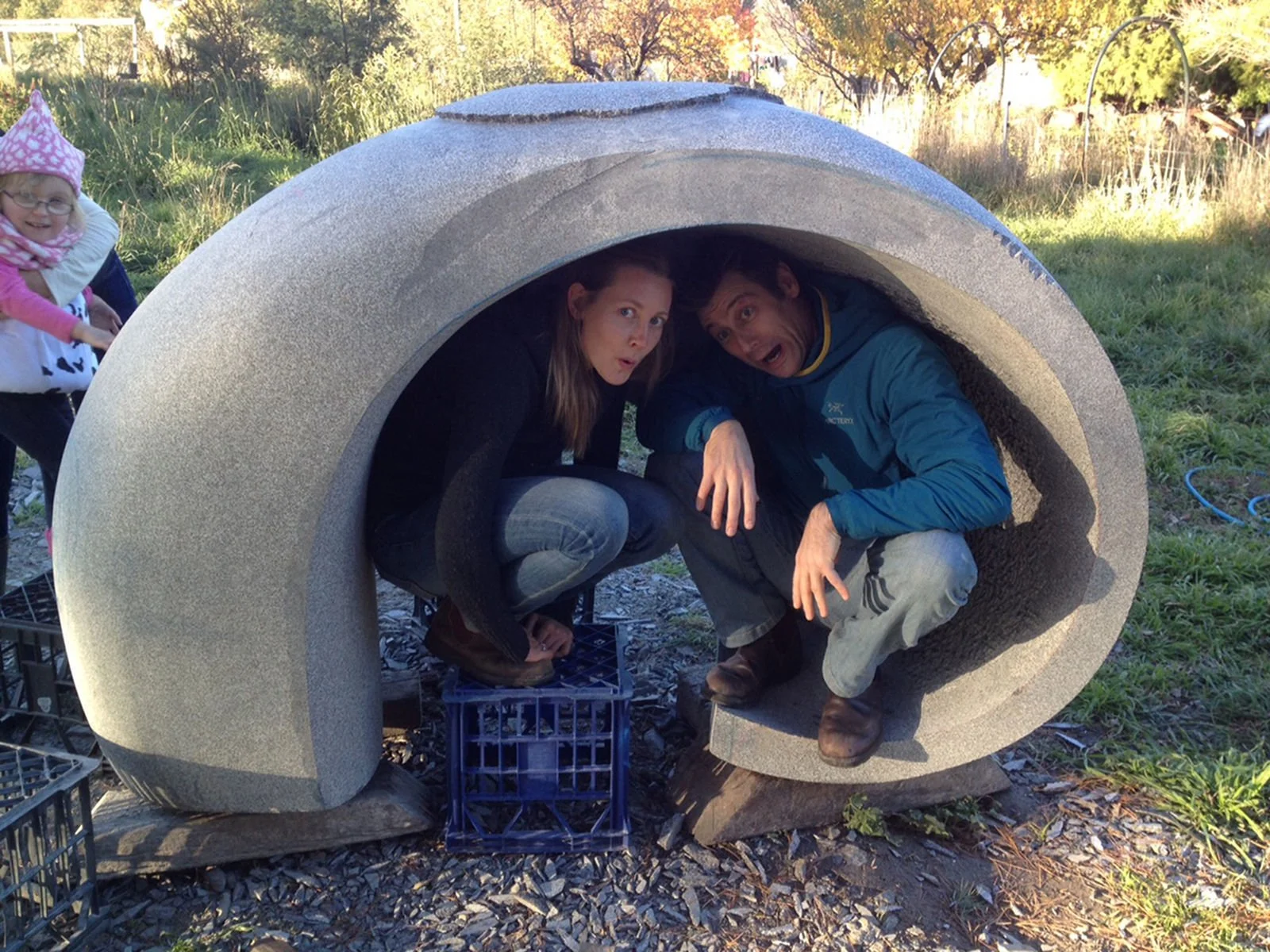 Two people crouching inside a large sculpture with surprised expressions, outdoors with grass and trees, and a small girl in pink on the left.