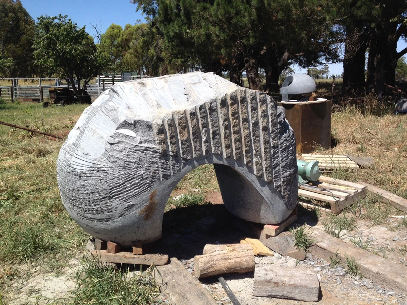 Large abstract stone sculpture outdoors in grassy field, with trees and equipment visible in the background.