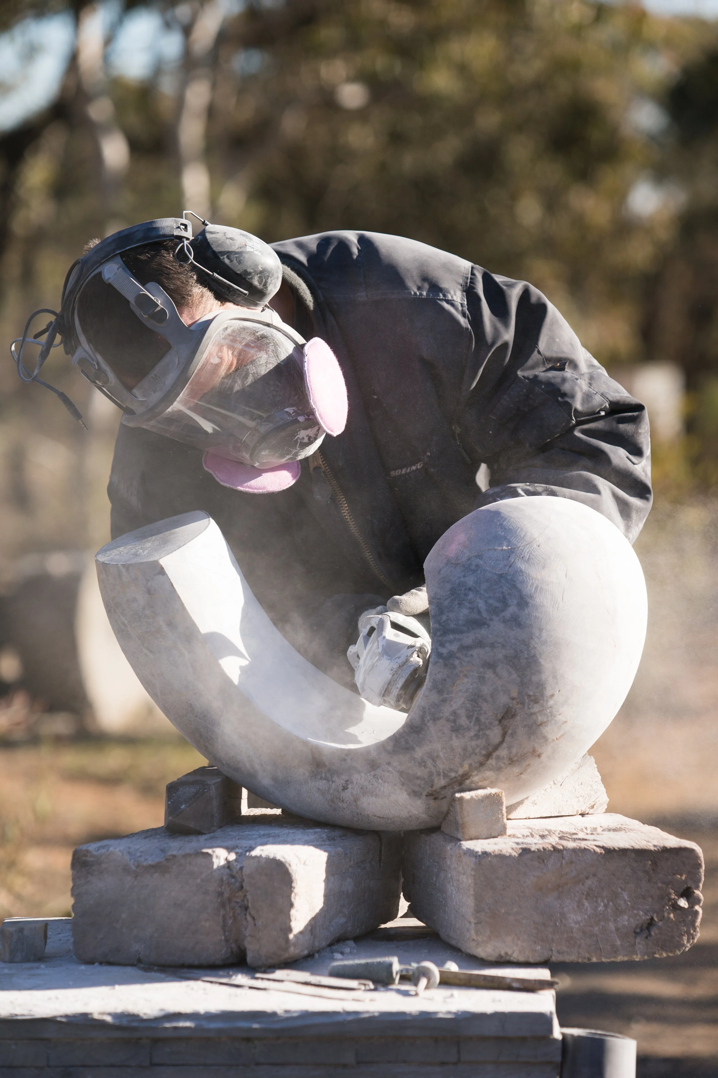 A person carving or sculpting a large, white marble sculpture outdoors, wearing a mask, protective goggles, and black jacket.
