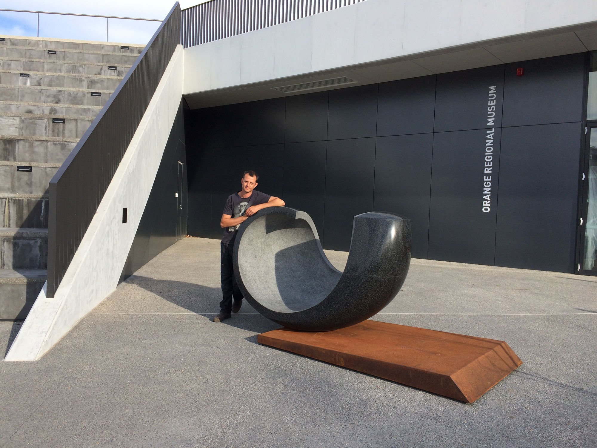 Man standing outdoors near a modern black sculpture resembling a large, curved, crescent shape, with the sign 'Orange Regional Museum' on a black wall behind him.