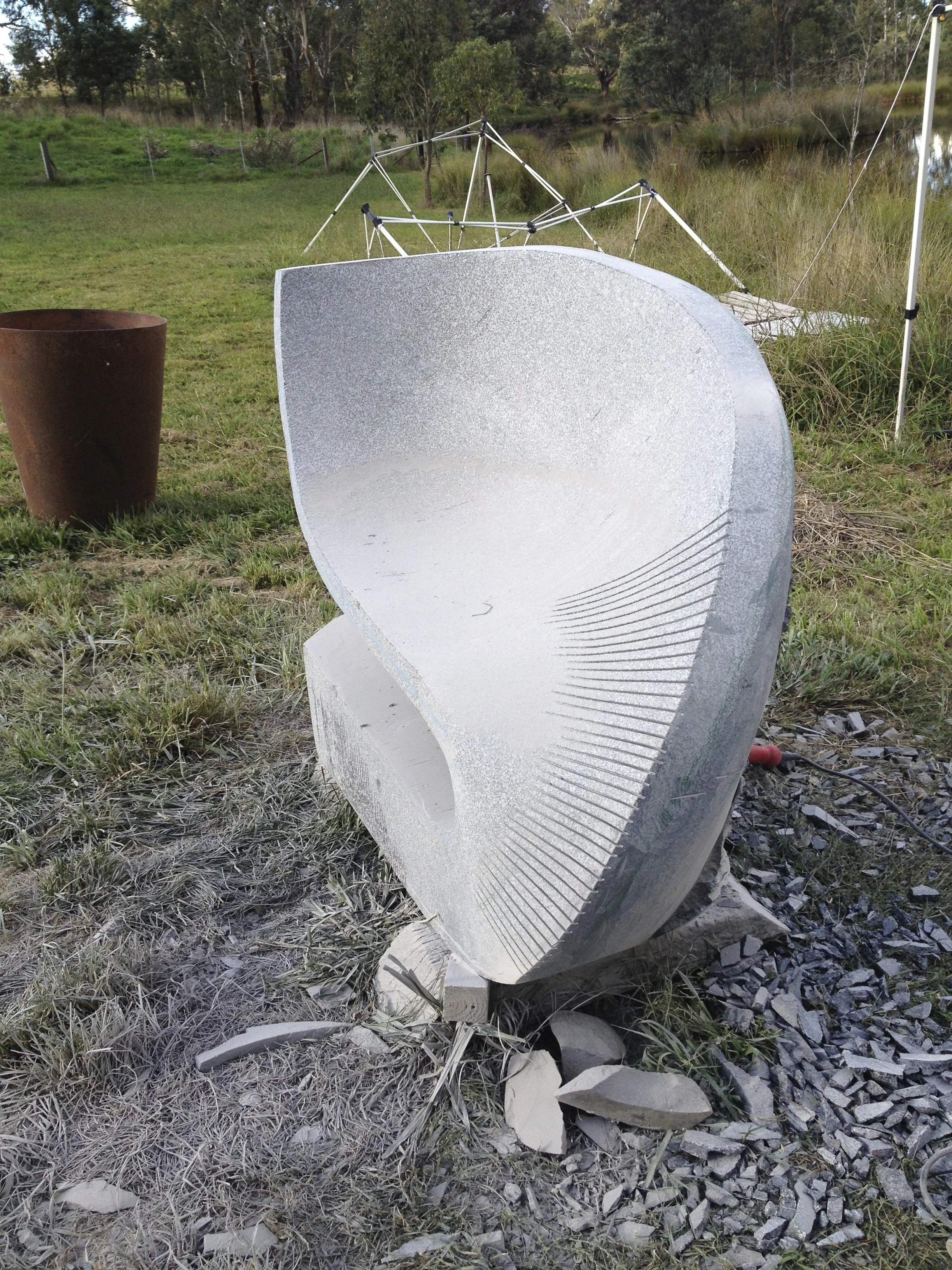 A stone sculpture outdoors in a grassy area with trees in the background and some broken pieces of stone on the ground.