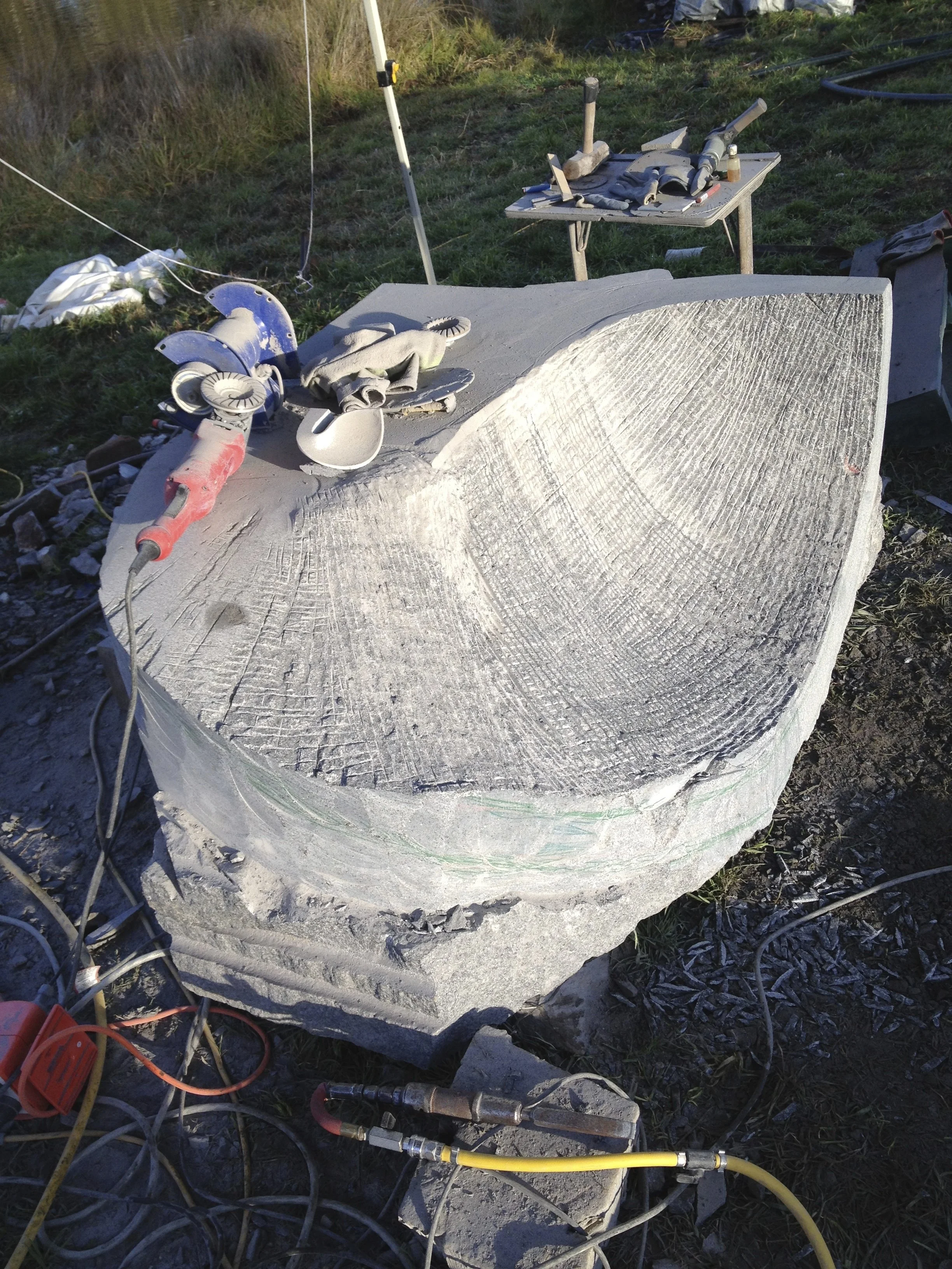 Close-up of a large stone block with numerous power tools, cables, and construction equipment scattered on and around it in an outdoor setting.