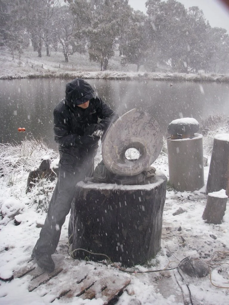 A person wearing dark clothing and a hood, standing outdoors in snowy weather, appears to be involved in sculpting or carving a large ice or stone piece on a tree stump. Snow covers the ground and the surrounding area, with trees and a lake in the ba