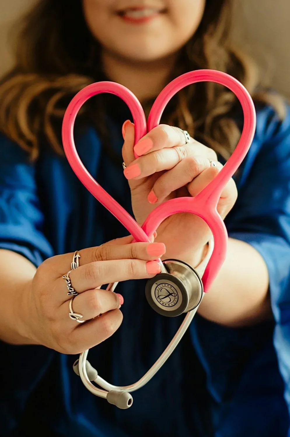 A woman holding a pink heart-shaped stethoscope in her hands, with her face slightly visible in the background, smiling.