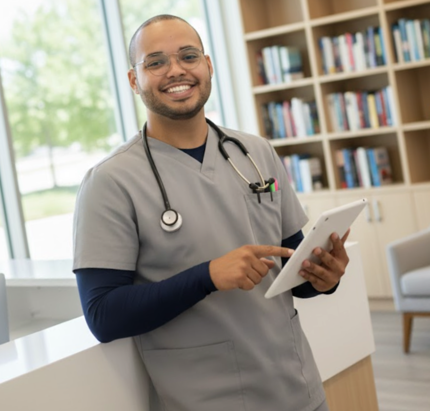 A smiling male healthcare professional in gray scrubs with a stethoscope around his neck, holding a tablet, standing in a medical office with bookshelves and a window.