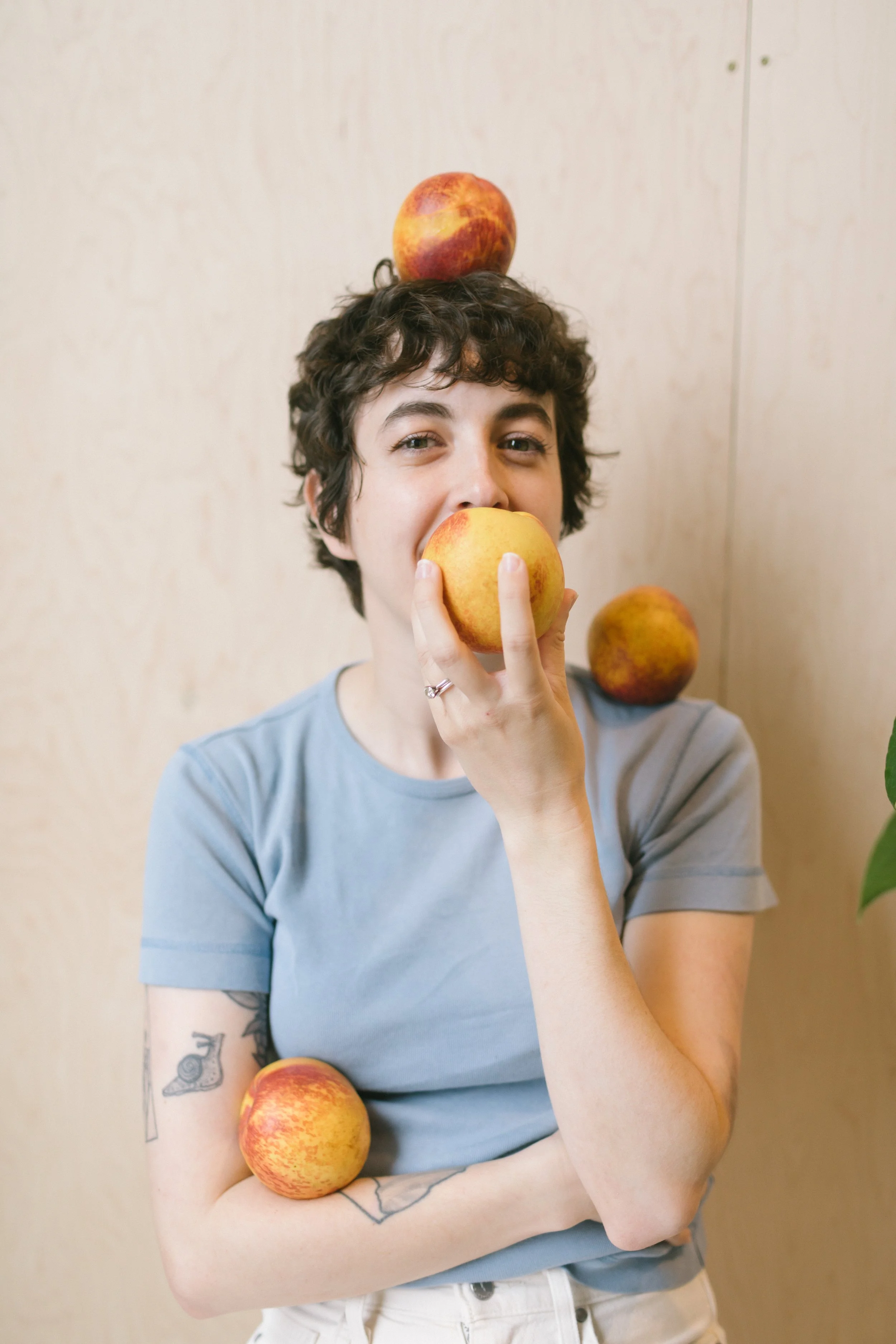 A person with short curly hair holding a large apple in front of their mouth, surrounded by three other apples, one on their head, one on their shoulder, and one against their arm, with a neutral background.