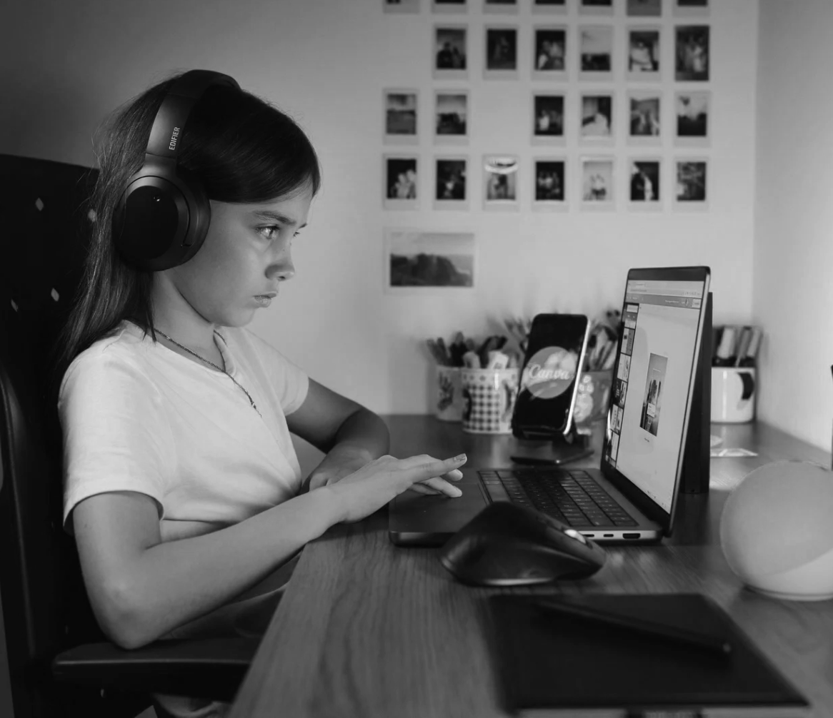A young girl with long hair wearing headphones working on a laptop at a desk. There is a smartphone, a mouse, and a notebook on the desk. The background has wall photos and containers with art supplies.