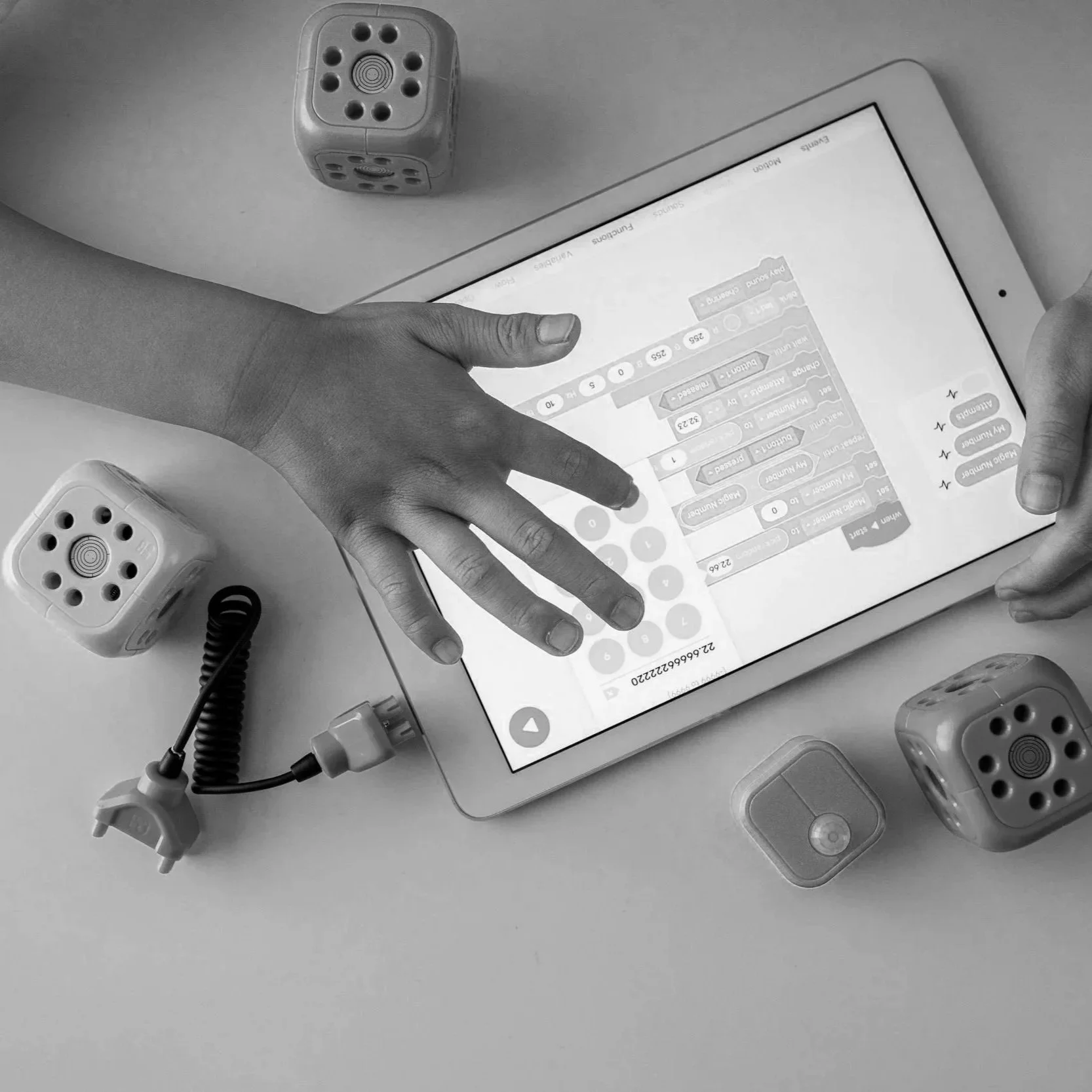 A person using a tablet with a block-based coding interface on the screen, surrounded by four dice, two of which have helicopter images, and a coiled charging cable.