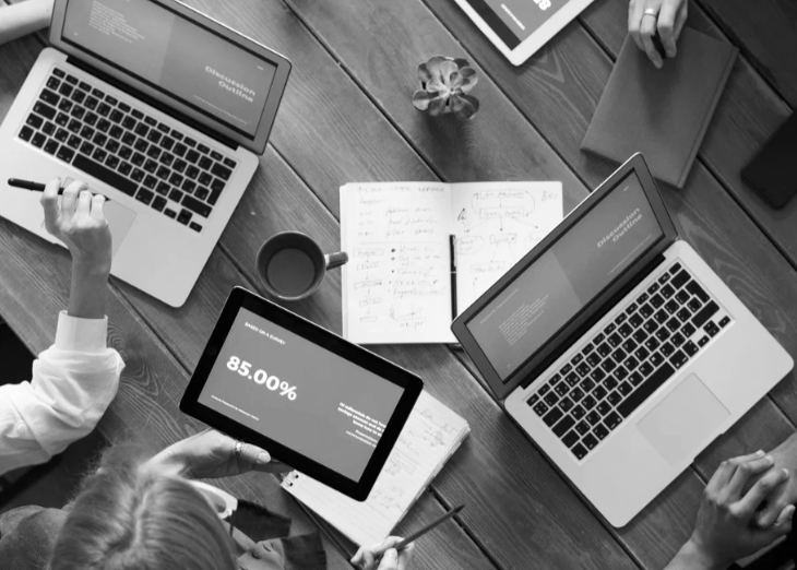 A top-down view of a meeting table with laptops, a tablet showing 85%, notebooks with handwritten notes, a cup of coffee, a potted plant, a tablet, and a smartphone.
