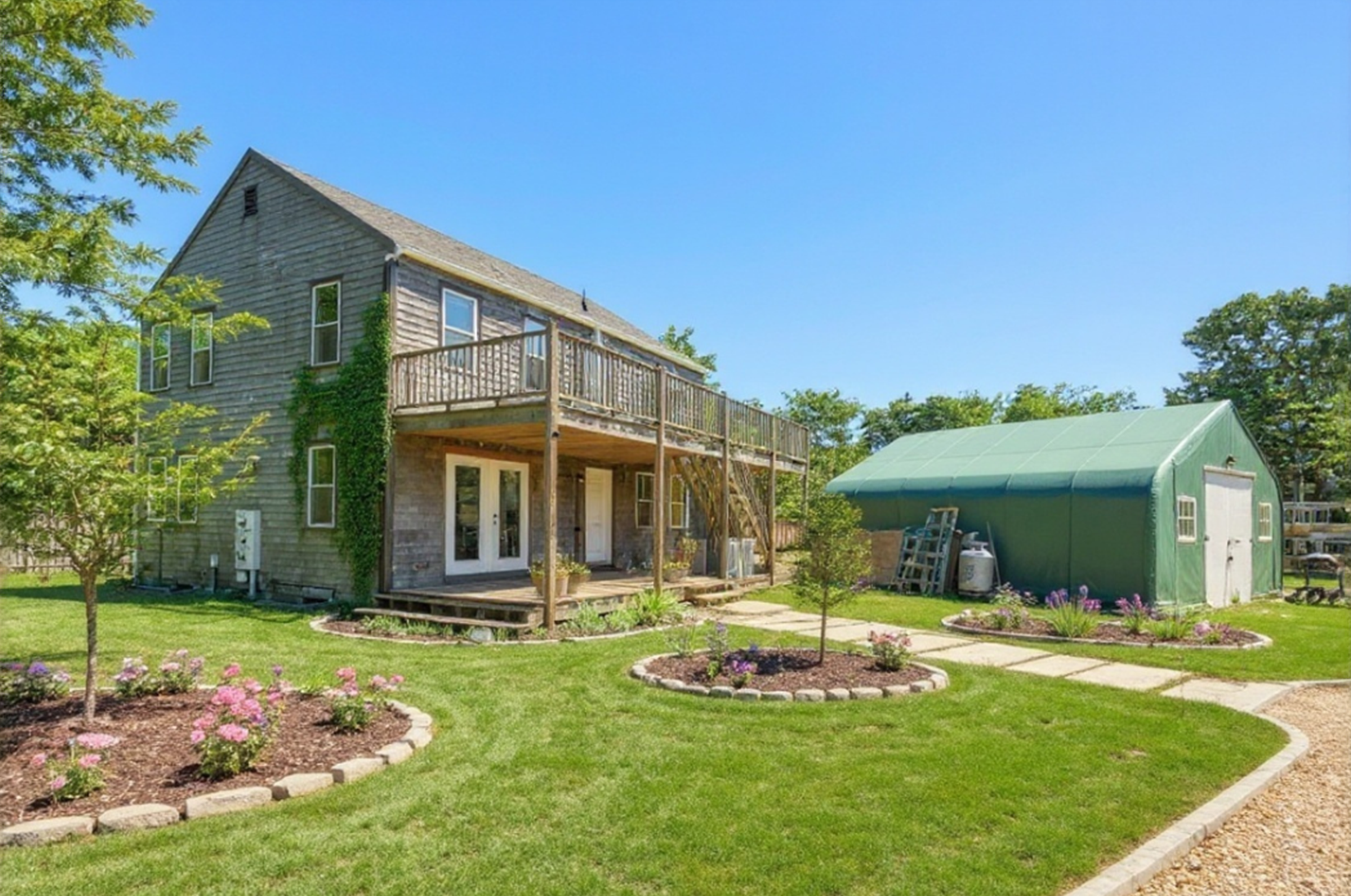 Backyard view of a Nantucket two-story home with balcony, colorful garden, and storage shed on a sunny day.