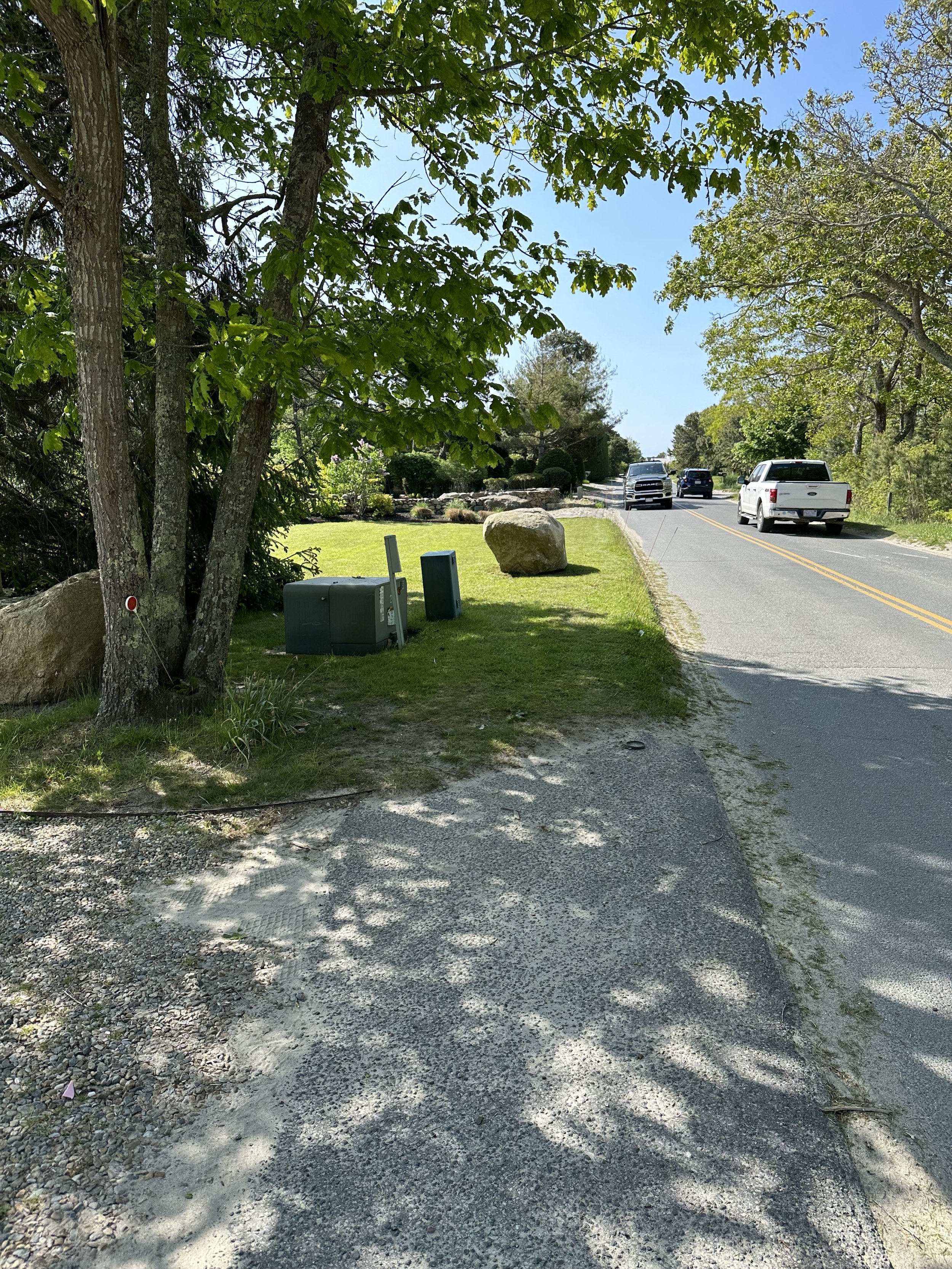 A roadside scene in a suburban area showing trees, large rocks, a well-maintained lawn, utility boxes, and a two-lane road with cars driving by on a sunny day.
