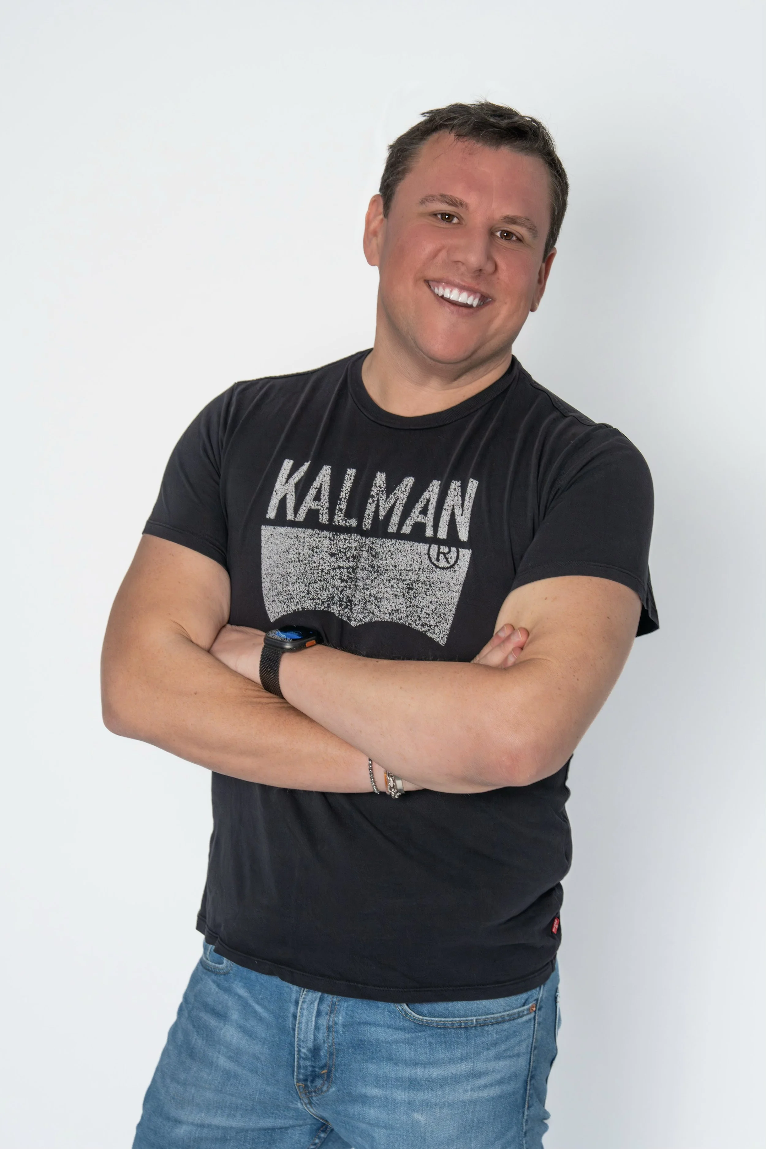 Sean Kalman Realtor from Nantucket smiling, crossing his arms, wearing a black T-shirt with 'KALMAN' printed on it, blue jeans, and a wristwatch, standing against a plain white background.
