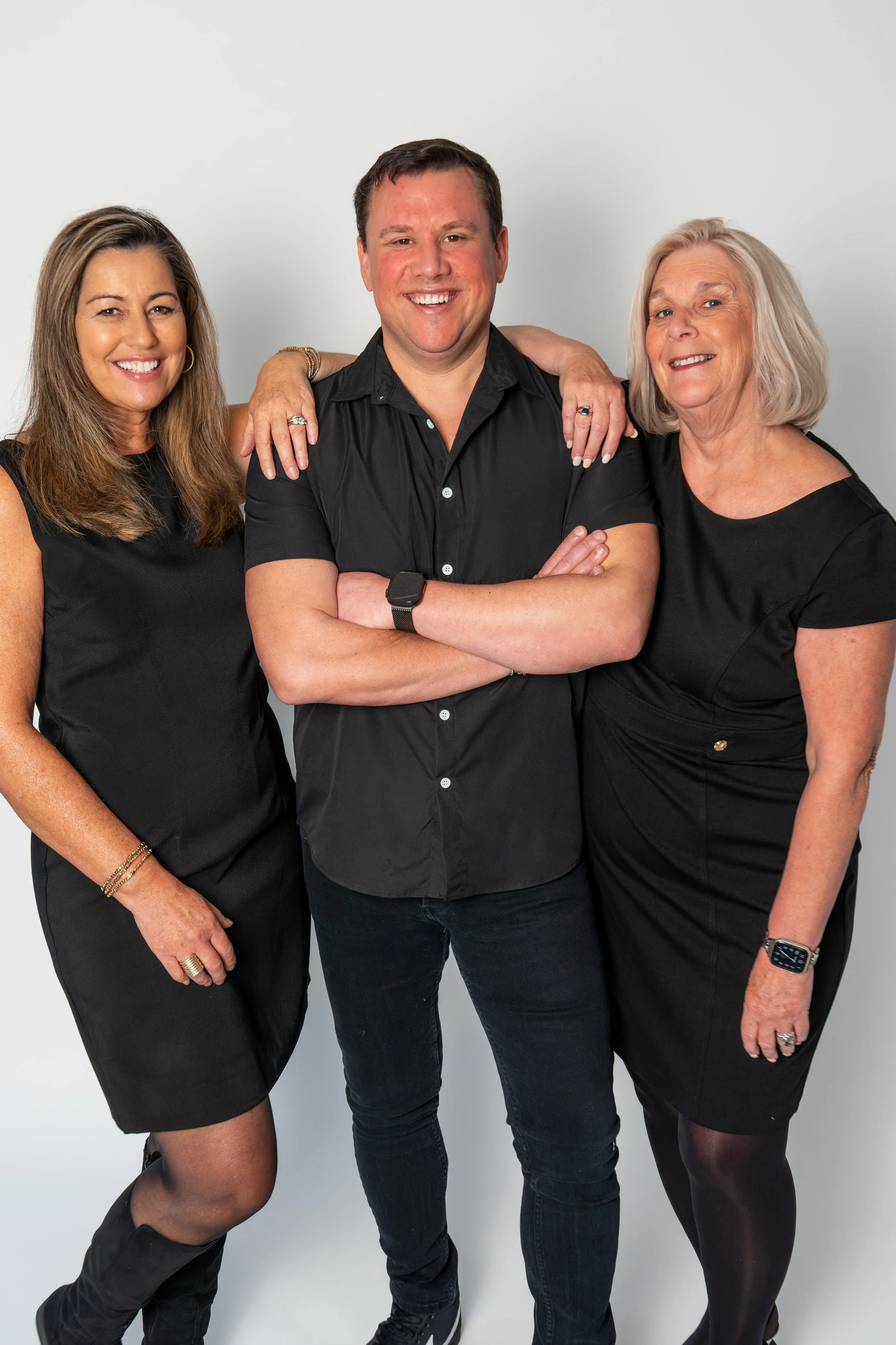 Three smiling people, two women and one man, dressed in black, standing against a plain white background. The women have shoulder-length hair, and the man has short dark hair.