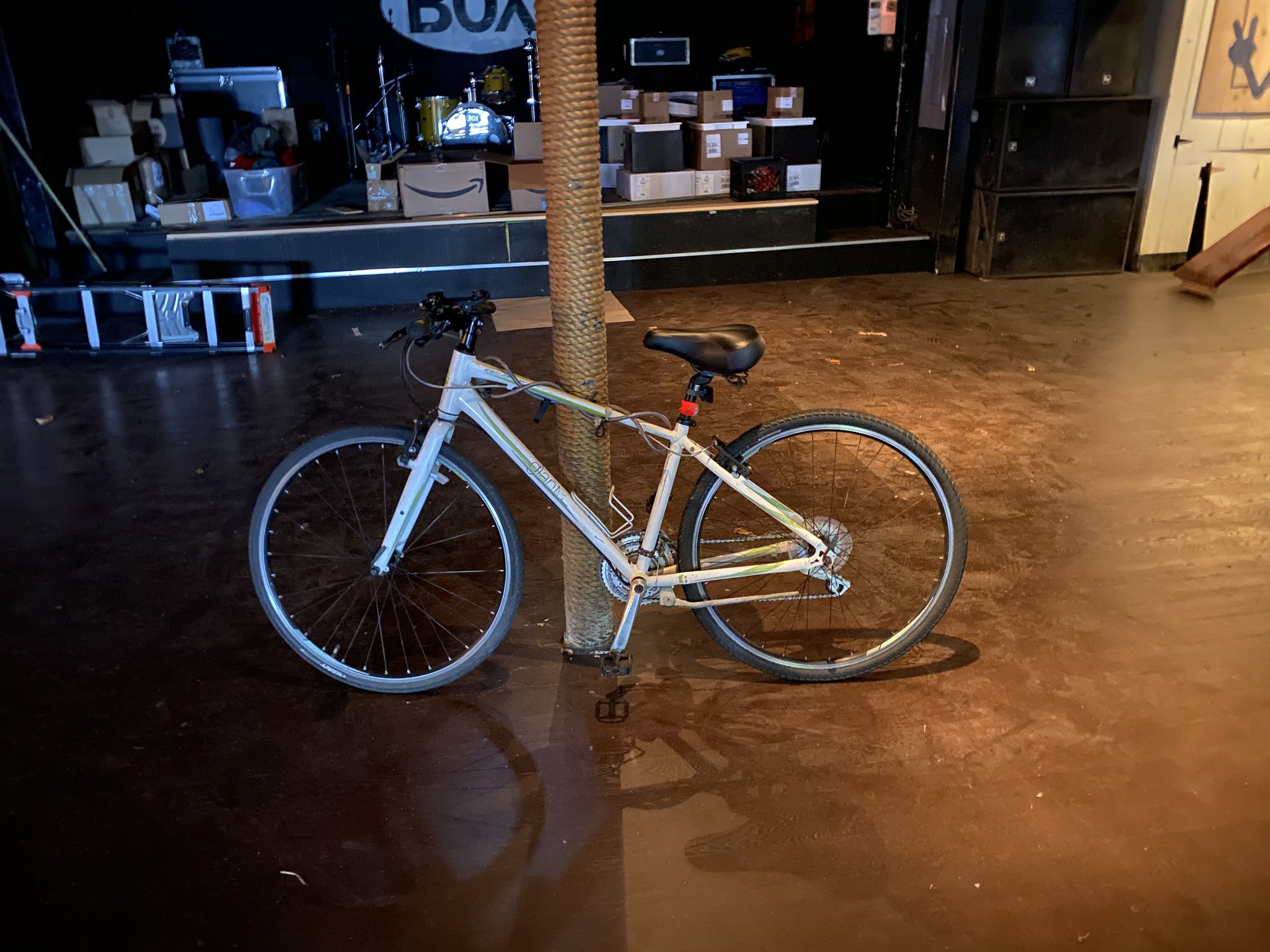 White mountain bicycle locked to a pole inside a dimly lit venue with a stage and boxes in the background.