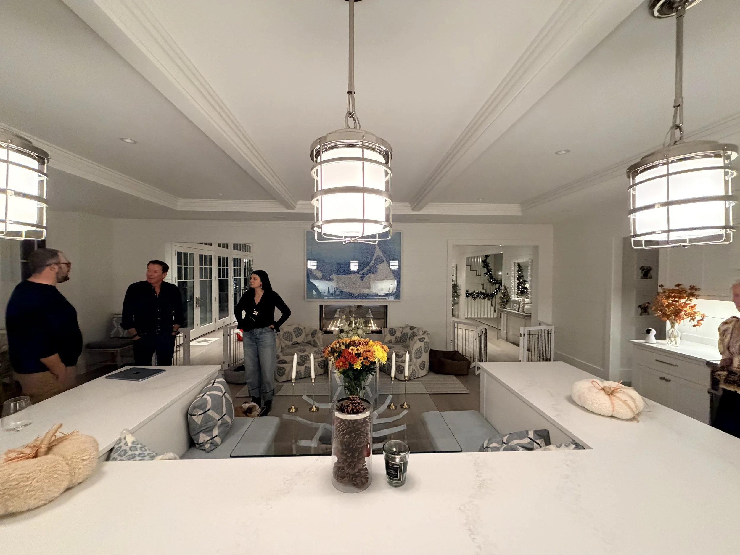 Modern house with wooden exterior in a mountainous landscape, with bushes and grass in the foreground, and two people visible through large glass window.