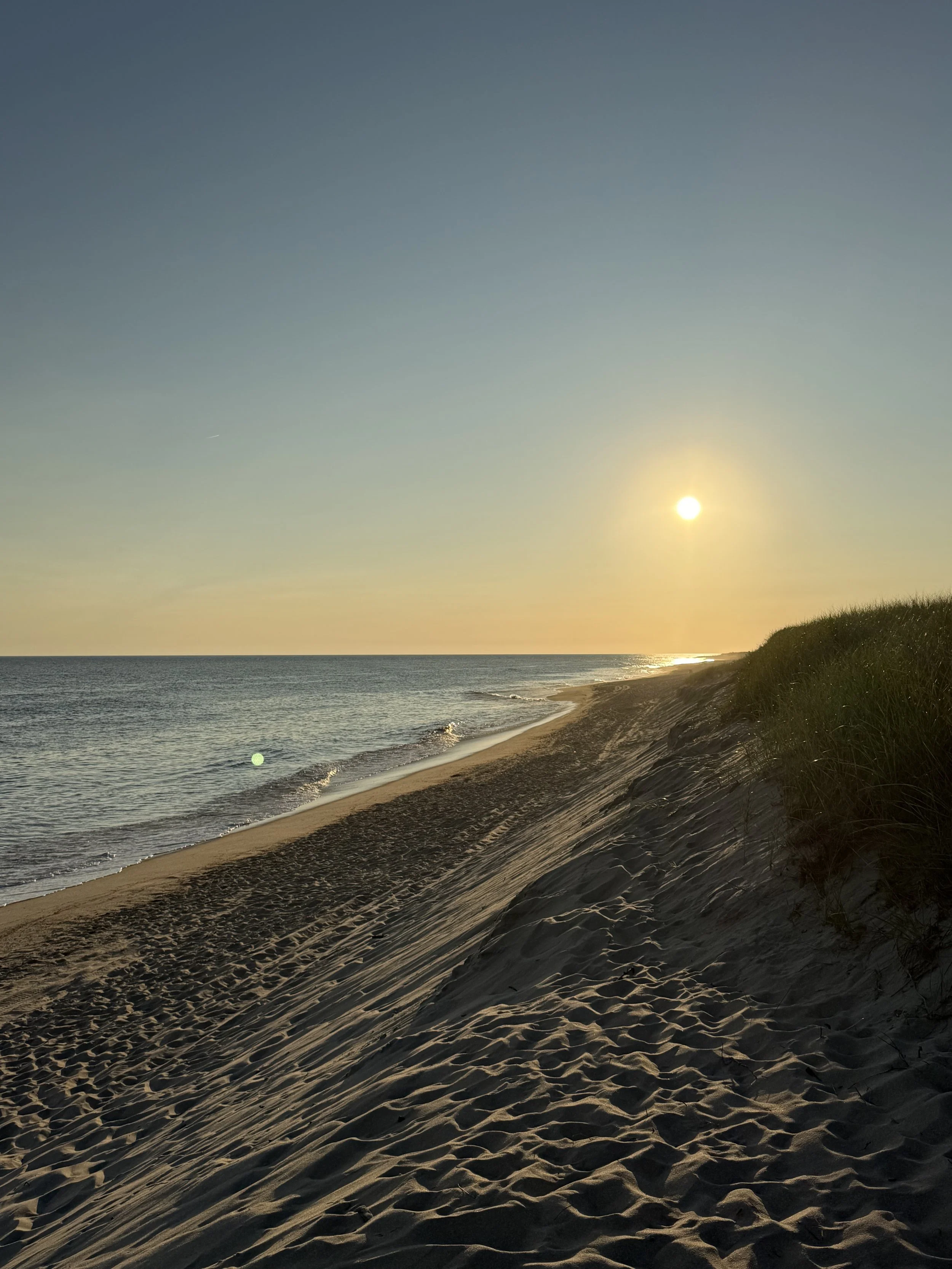 Sunset over a sandy beach with grassy dunes on the right, gentle waves hitting the shore, and a clear sky.