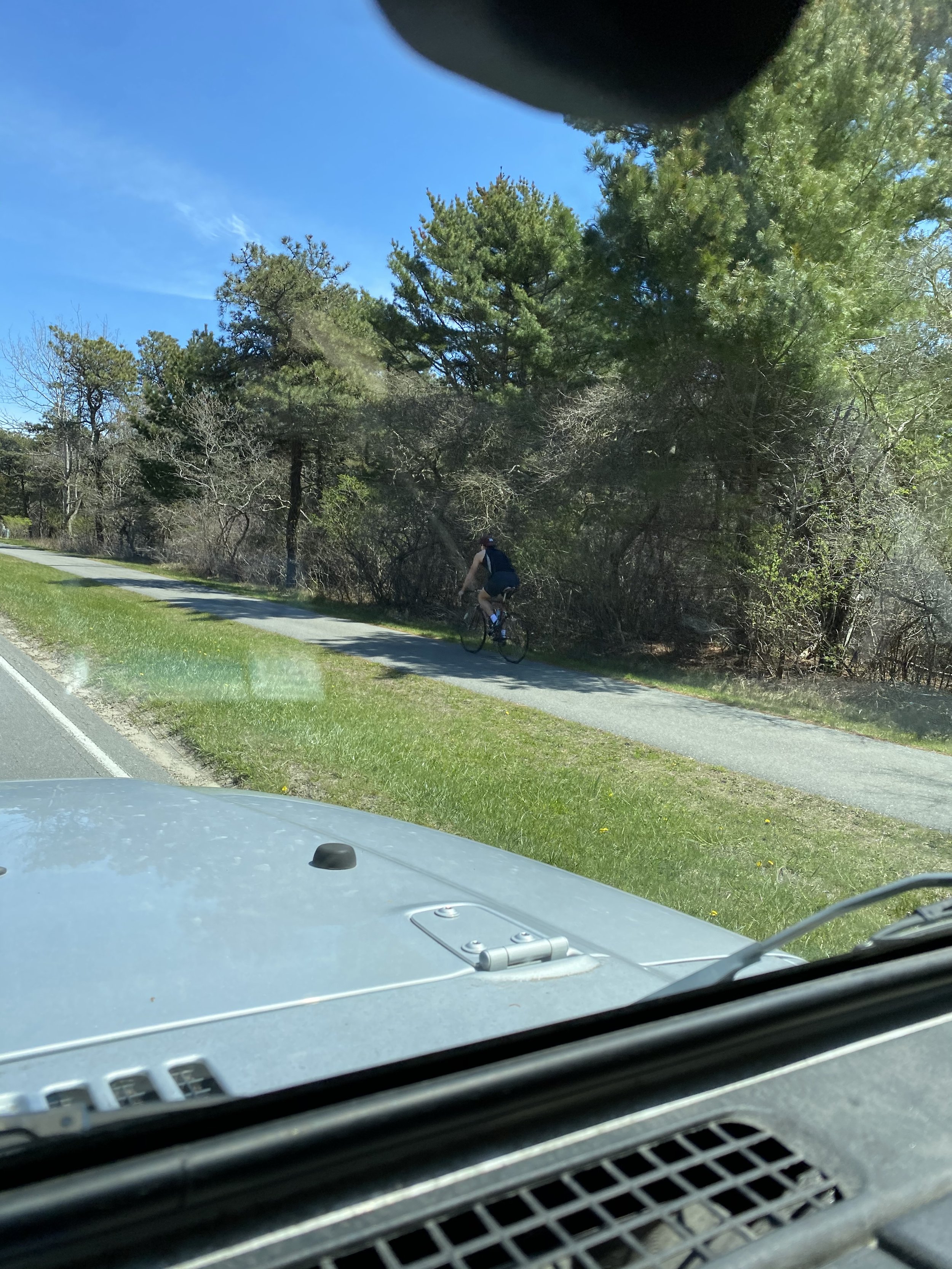 A person riding a bicycle on a paved trail beside a grassy area and trees under a clear blue sky, viewed from inside a vehicle.