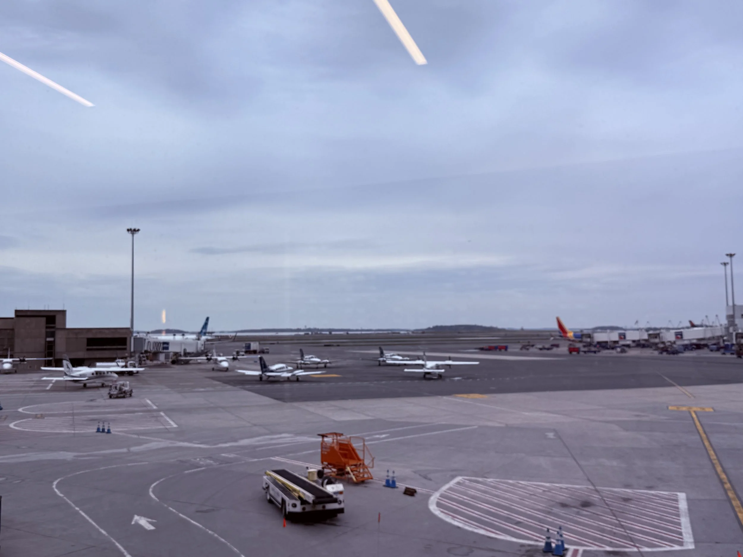View of an airport tarmac with several small airplanes parked near terminal gates, connecting jet bridges, and ground service vehicles under an overcast sky.