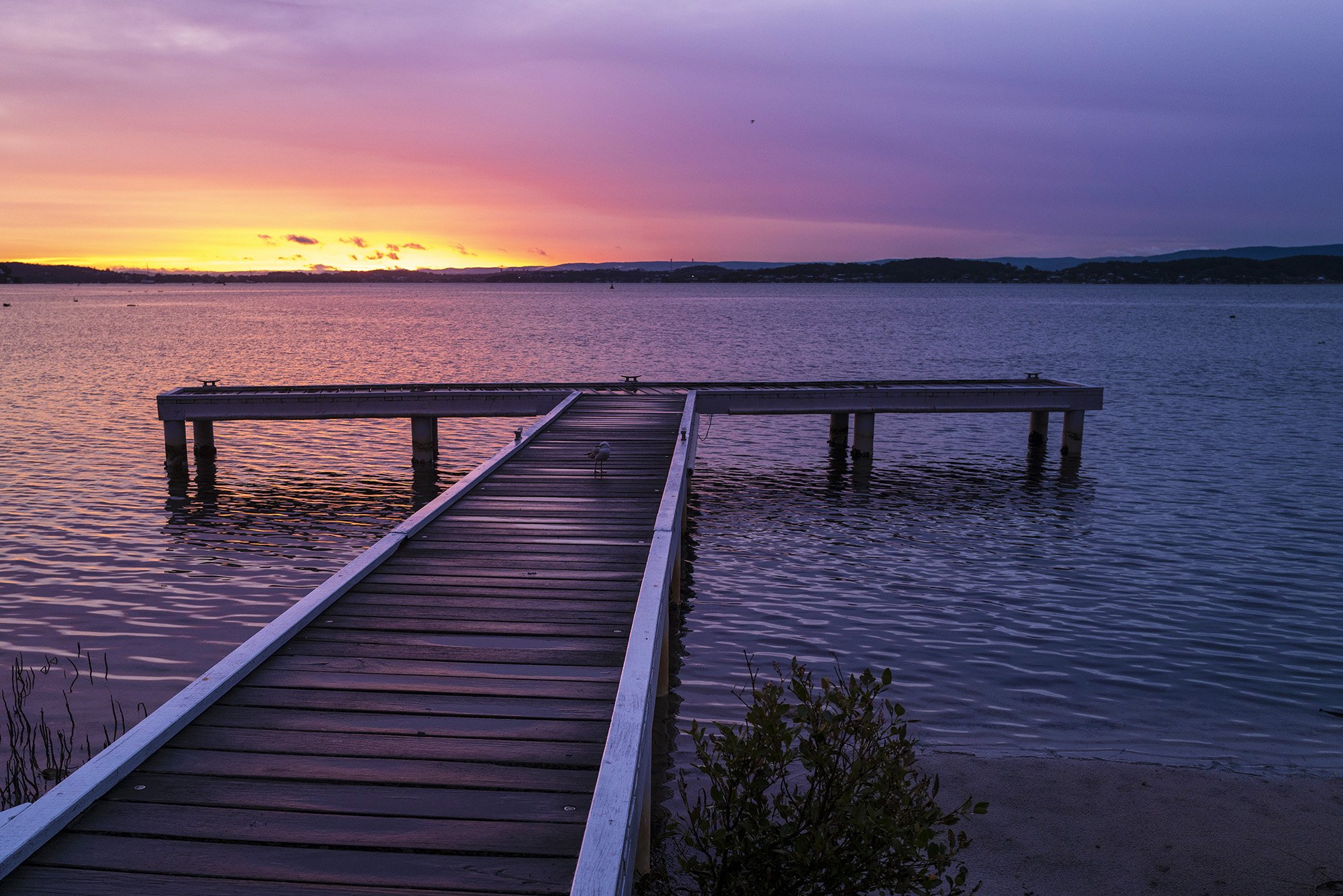 A wooden dock extending into a calm body of water during sunset, with colorful orange, pink, and purple sky and distant hills.
