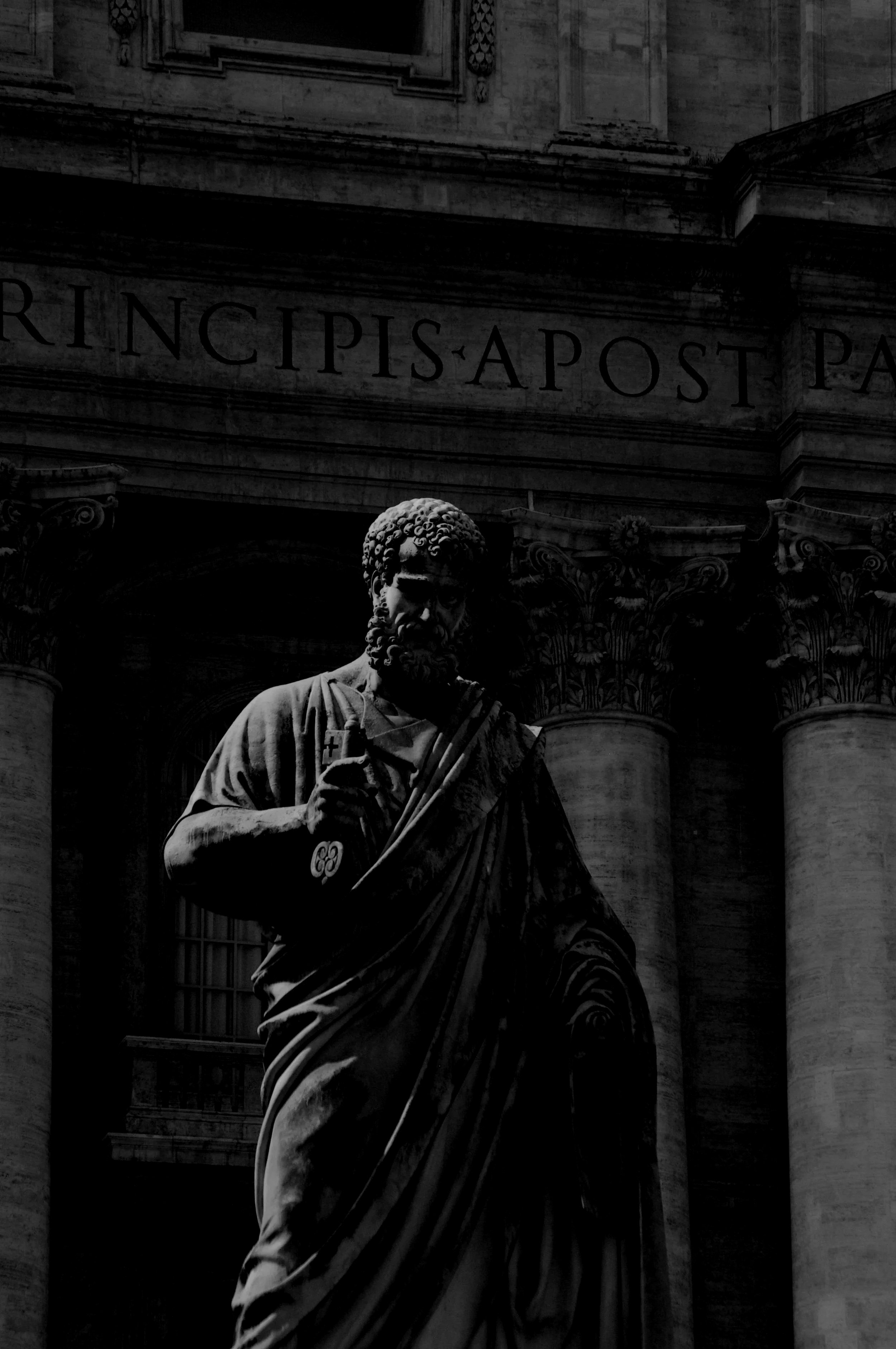 Black and white photograph of a classical statue of a bearded man holding a cross, in front of a building with columns and Latin inscriptions.