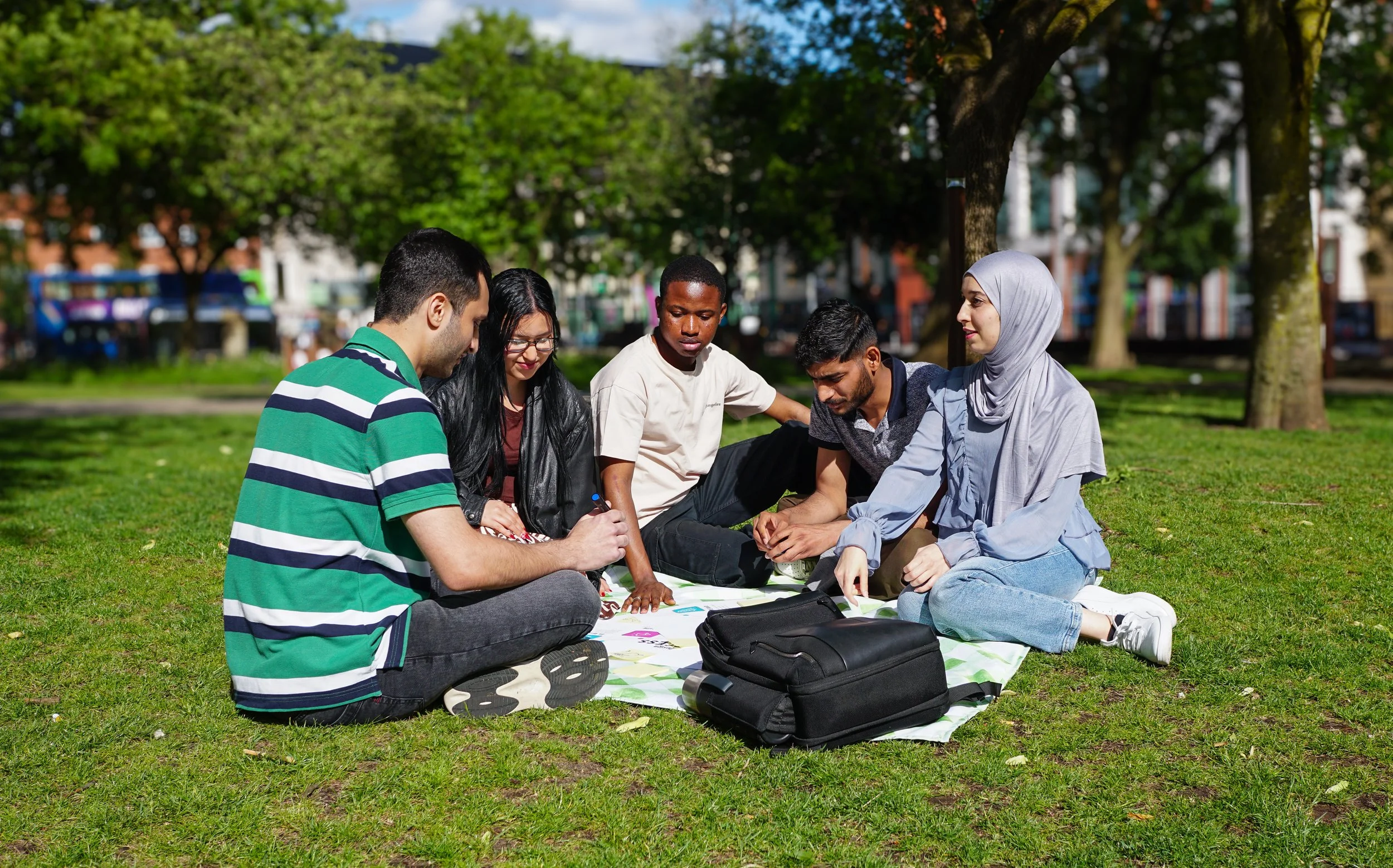 Group of five diverse young adults sitting on a picnic blanket in a park, engaged in a board game, with trees, grass, and buildings in the background.