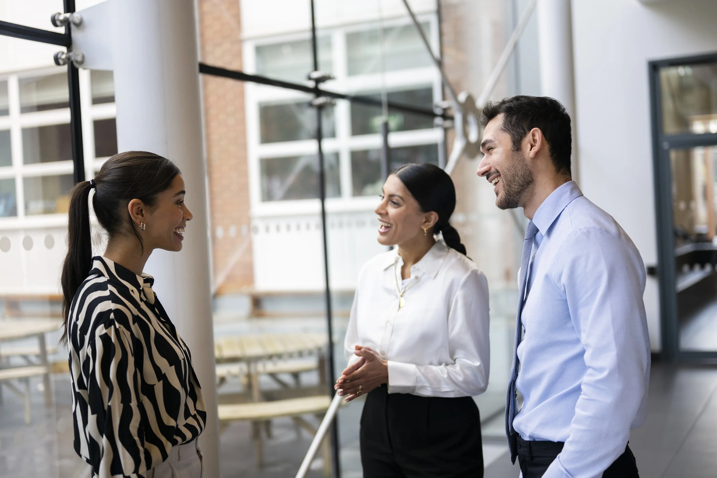 Three professionals, two women and one man, engaged in a friendly conversation in a modern office space with large windows and a glass wall.