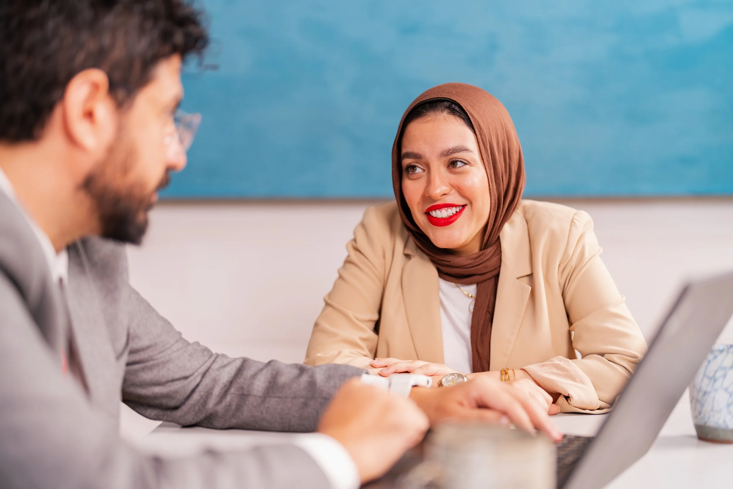 A woman wearing a hijab and beige blazer smiling at a man with glasses and beard, sitting at a desk with a laptop.