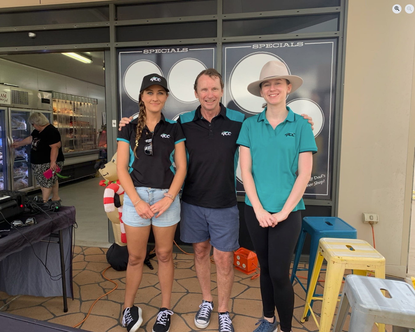 Three people smiling and posing for a photo inside a building near a booth or sign that says 'SPECIALS.' The two women are wearing black and teal shirts with a logo, and the man is wearing a black polo shirt with the same logo. The woman on the right has a wide-brimmed hat. There are stools and electronic equipment around them.