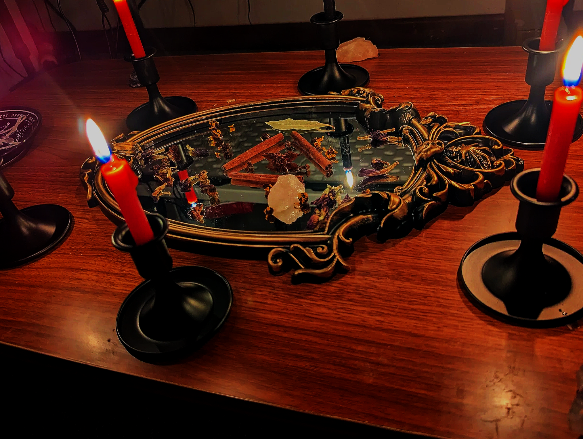 A wooden dining table with a decorative black and gold-framed mirror in the center, surrounded by five black candlestick holders with red candles. The mirror reflects some cinnamon sticks, dried herbs, and a rock salt crystal, with the candles lit.