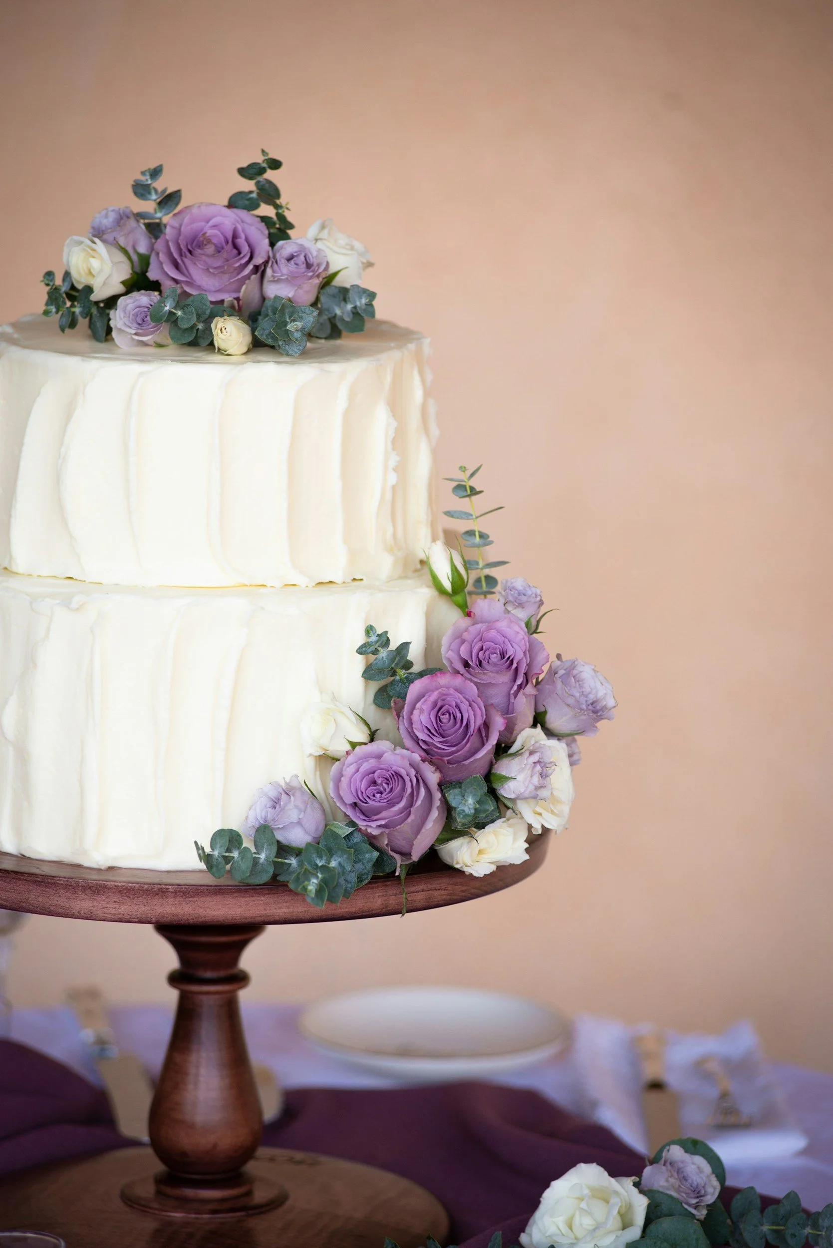 Elegant two-tier wedding cake decorated with purple and white roses and greenery on a wooden cake stand.