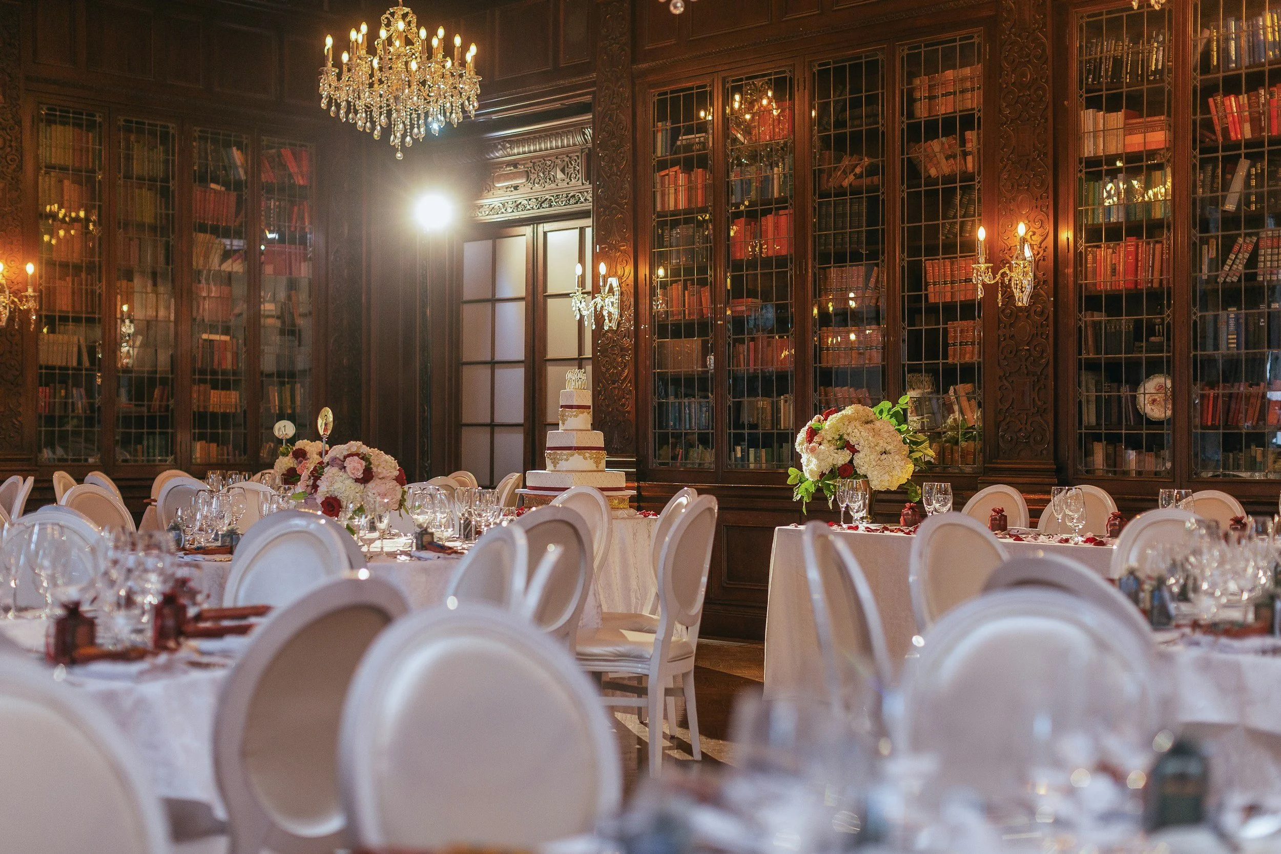 Elegant wedding reception setup in a grand, wood-paneled library room with a chandelier, bookshelves, a wedding cake, floral centerpieces, and neatly arranged dining tables.
