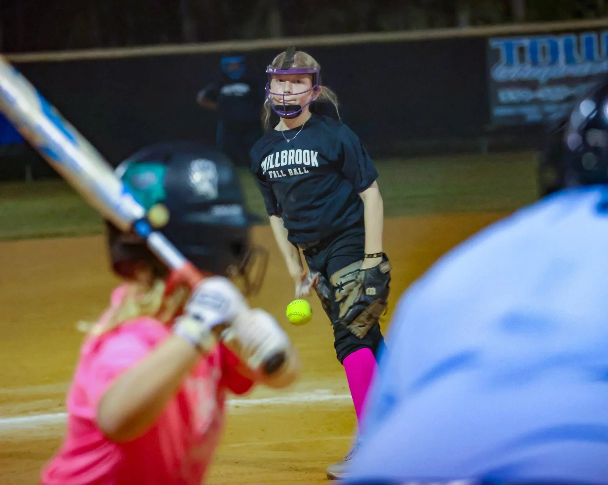 A young girl in pink softball gear swinging a bat, with a softball pitcher in the background on a softball field at night.