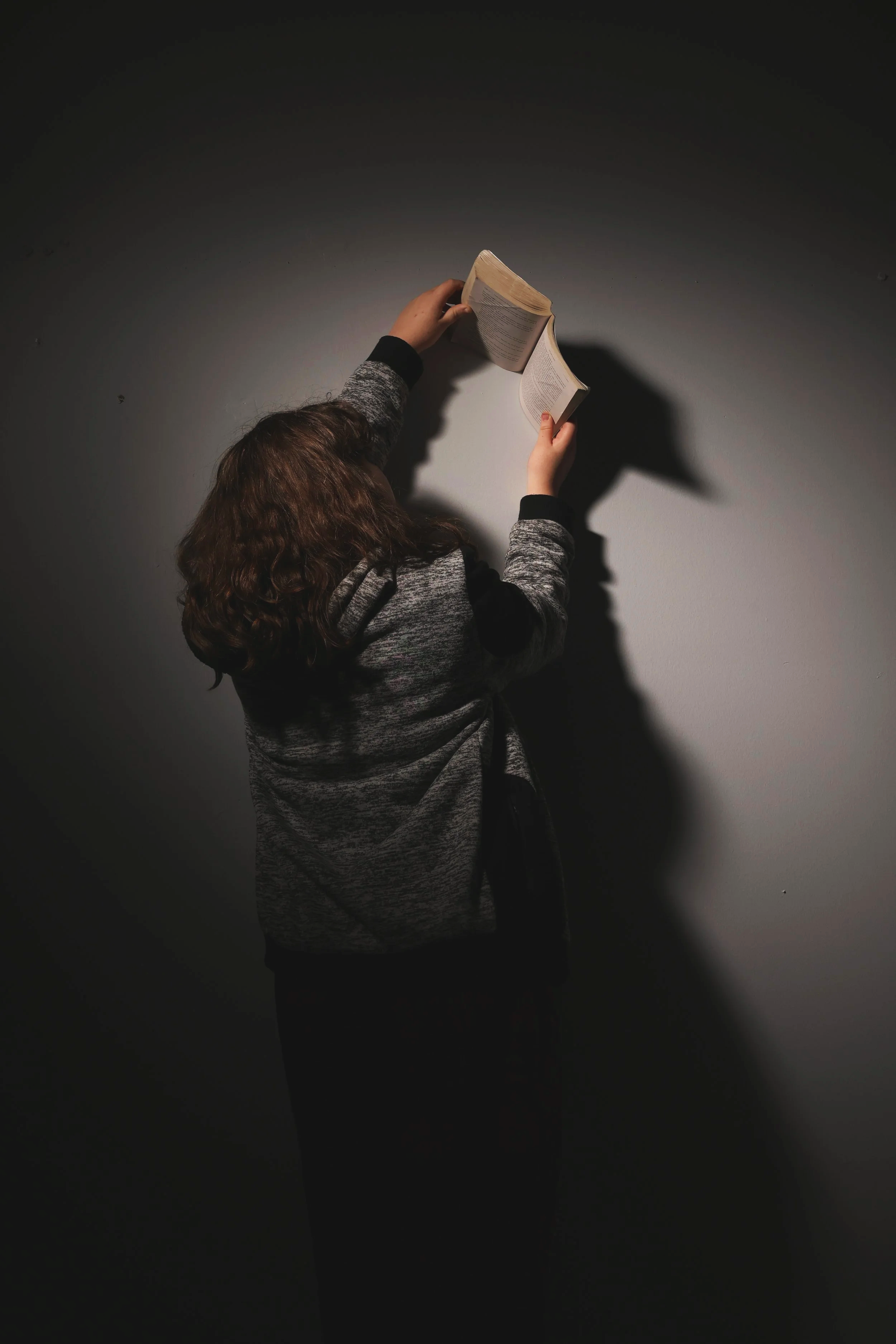 Person with curly hair wearing a gray sweater reading a book against a dark gray wall, casting a shadow.