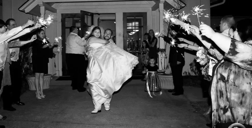 Joyful bride in wedding gown being carried by groom, surrounded by friends and family holding sparklers