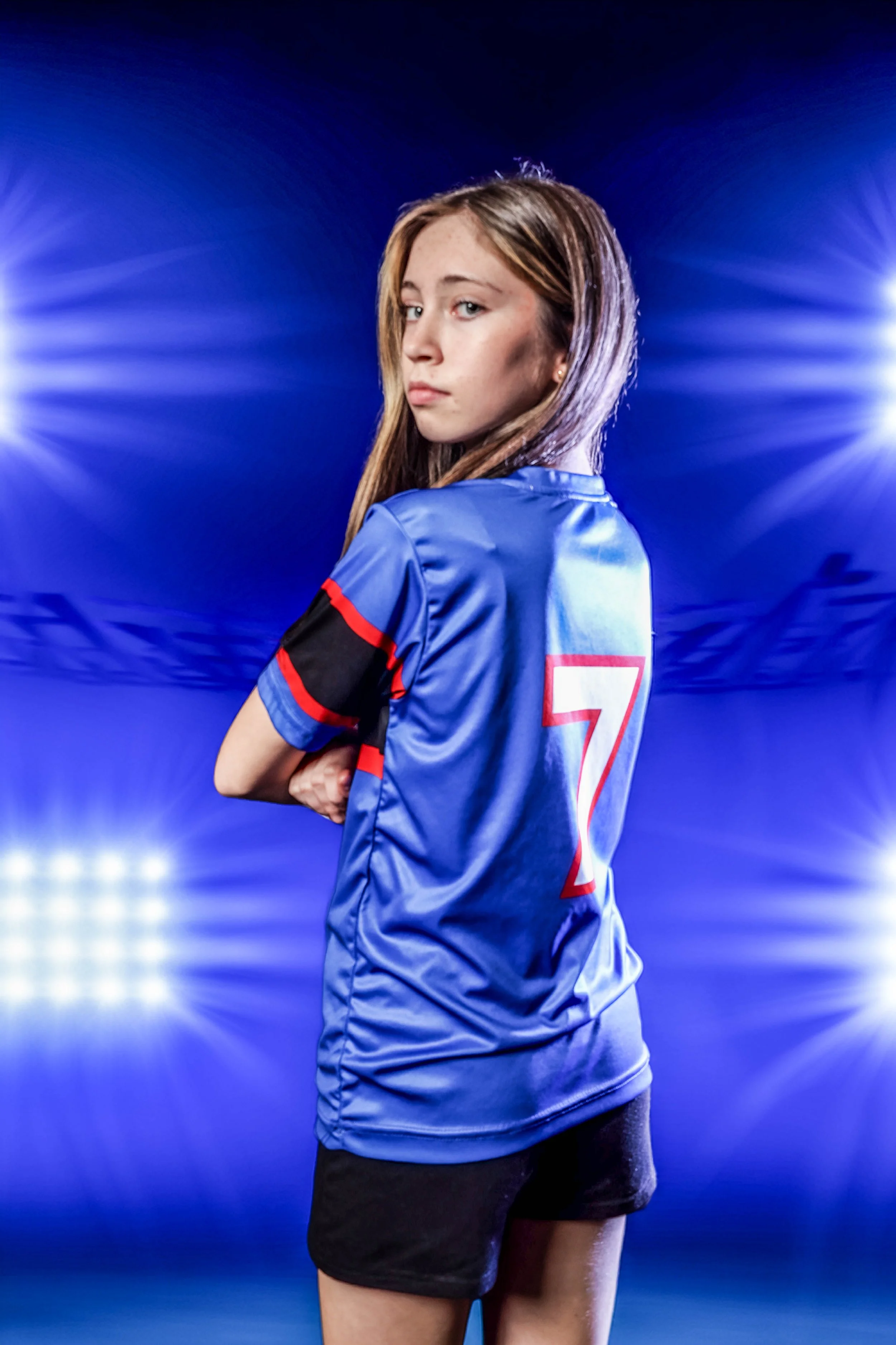 Young female soccer player wearing a blue jersey with the number 7, standing with arms crossed on a lit sports field at night with stadium lights in background.