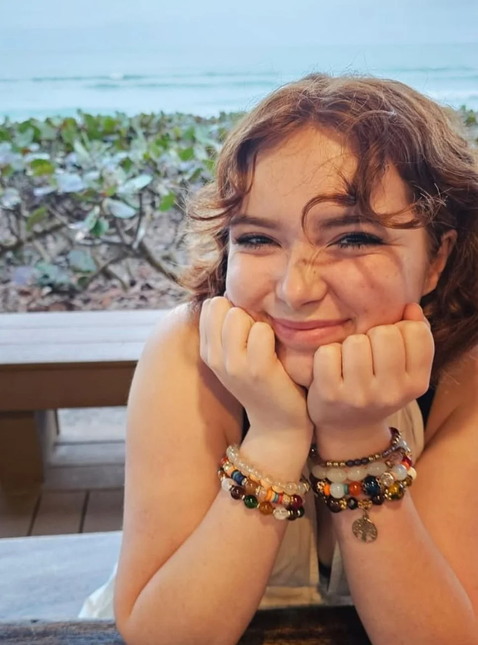 A young woman with curly red hair is smiling as she rests her chin on her hands, sitting at a table near the beach with greenery and ocean in the background.