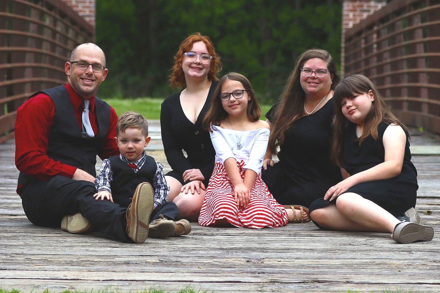 A family of six sitting on a wooden bridge outdoors on a cloudy day.