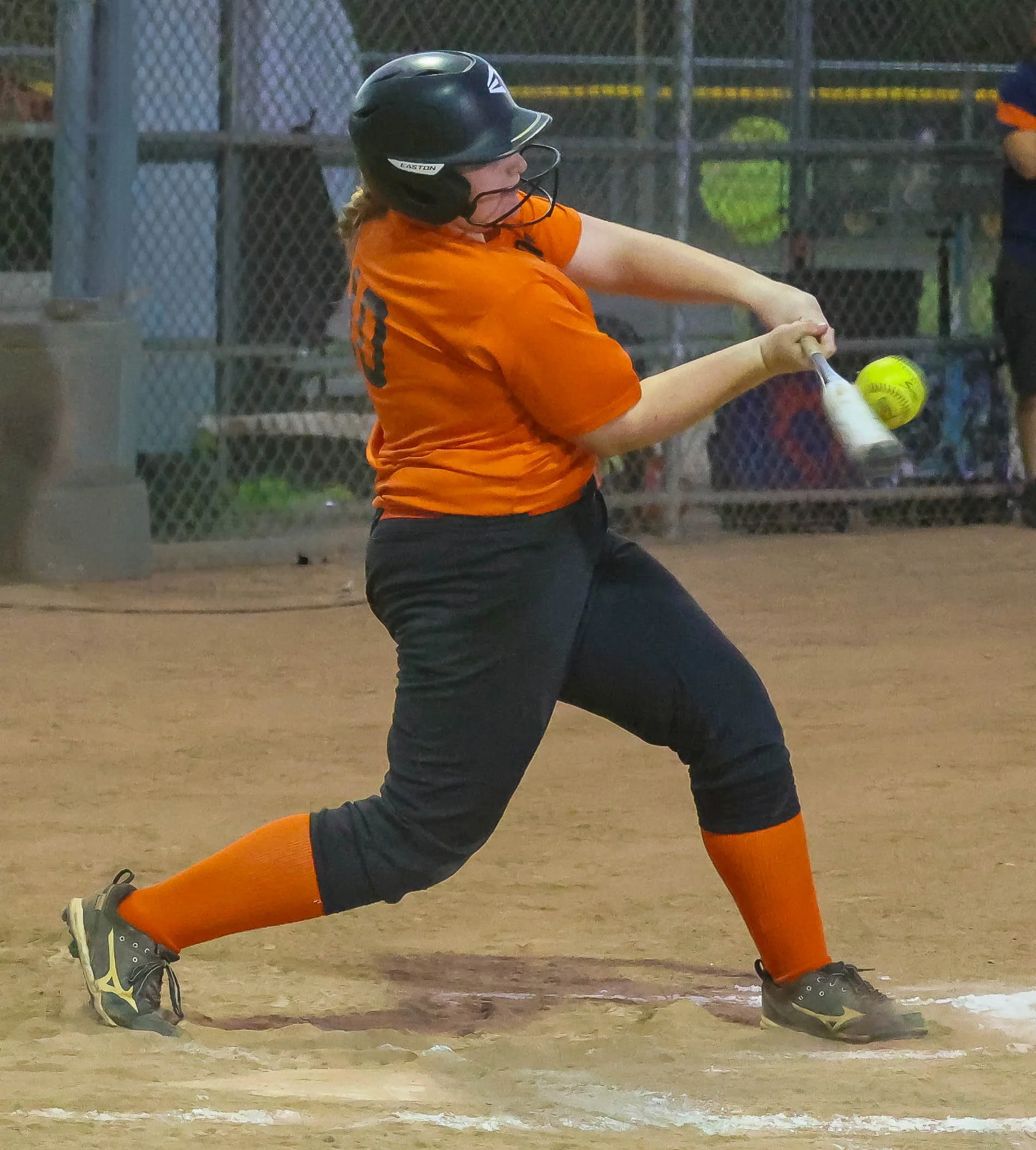 A softball athlete in an orange shirt, black pants, and orange socks is swinging a softball bat at a pitched softball on a softball field, wearing a black helmet.