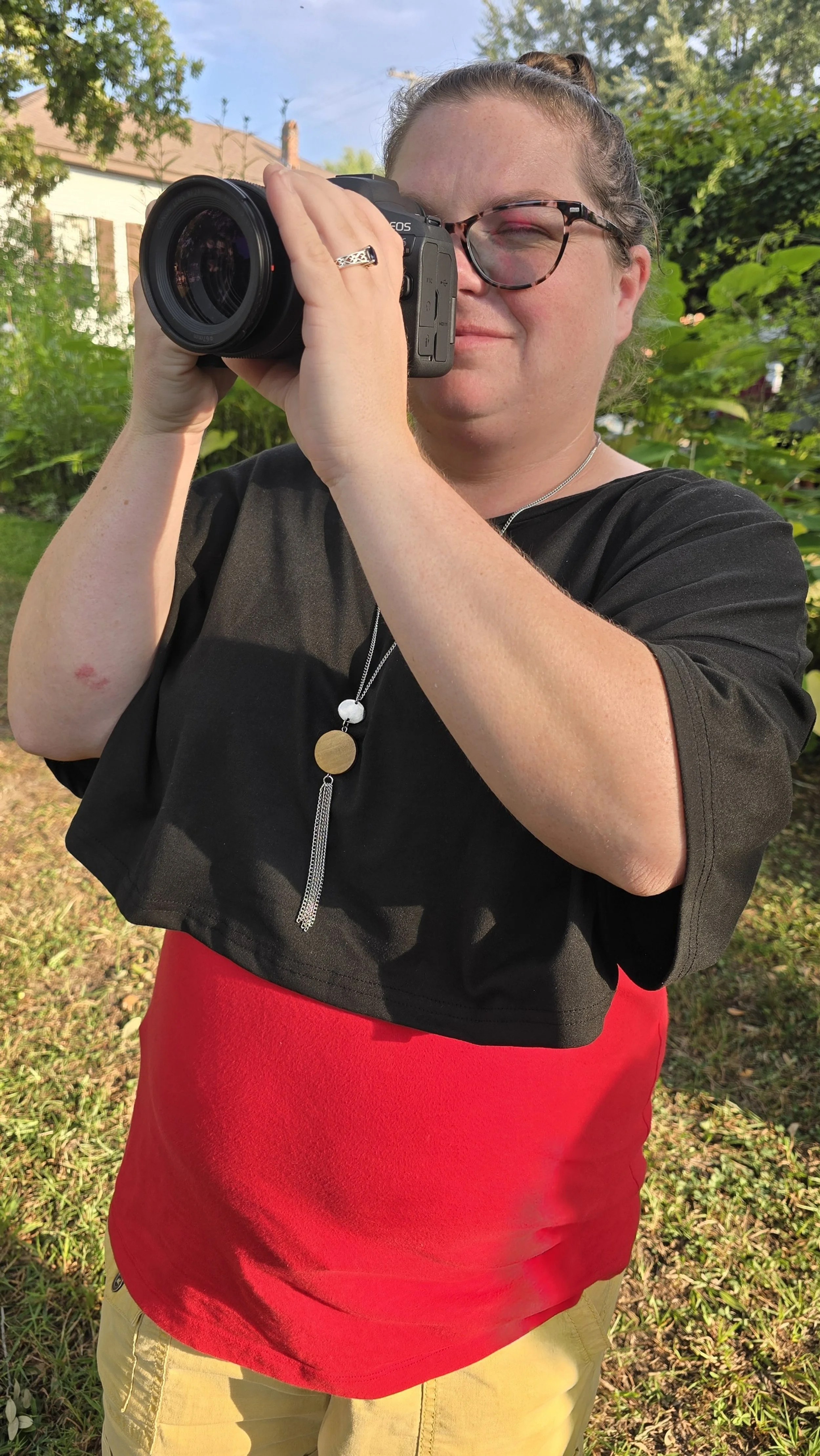 A woman with glasses and a ponytail is holding a camera up to her face, taking a photo outdoors in a garden or yard area with green foliage and a building in the background.